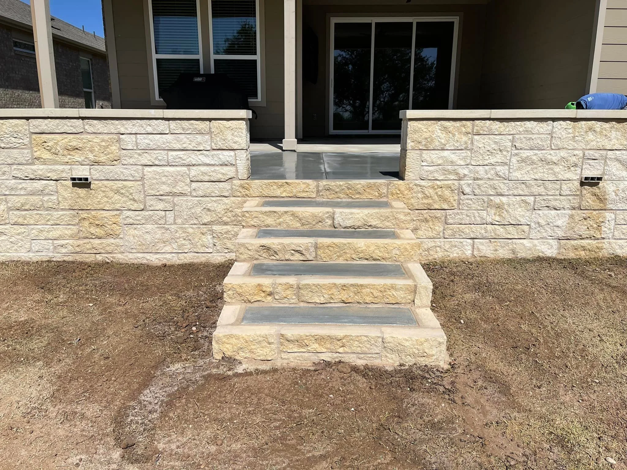Stone steps leading to a backyard patio, with a stone wall on either side, in front of a house with sliding glass doors and windows, and a dirt yard in the foreground.