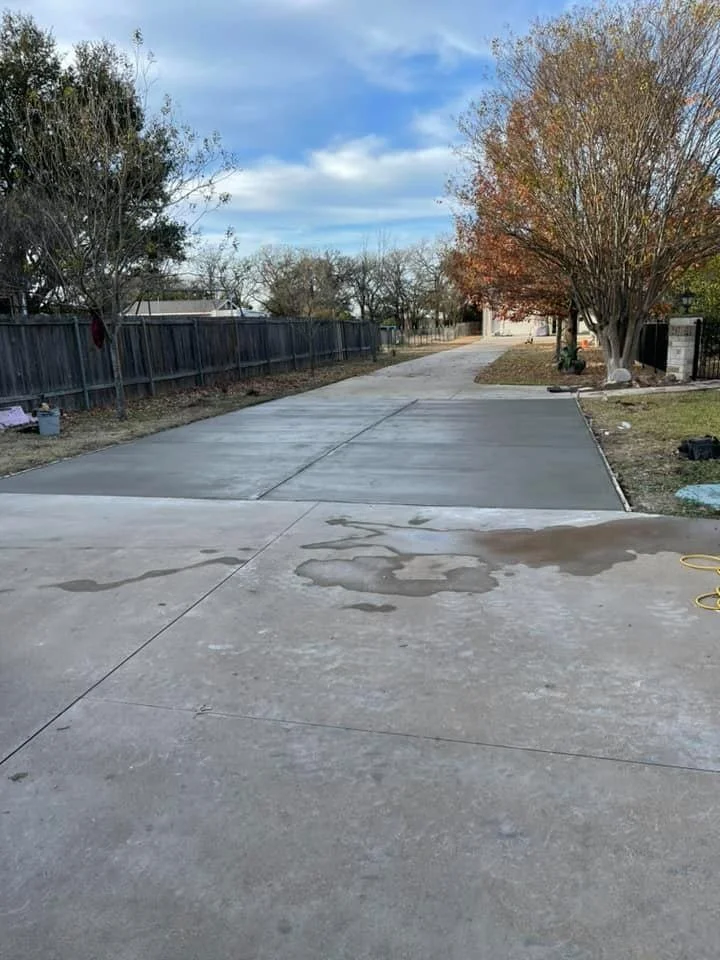 A driveway in a suburban neighborhood with parts of the concrete freshly poured, some wet spots, and leafless trees on either side.