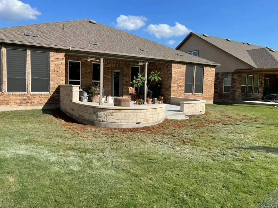 Backyard with a small stone wall patio, potted plants, and a covered area. Brick house with multiple windows in the background.
