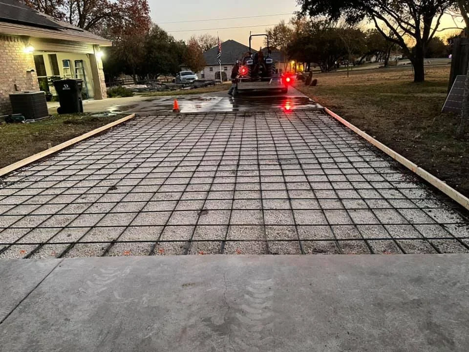 A residential driveway under construction, with a grid of rebar laid out over gravel, and a construction vehicle at the back with orange safety cones nearby, during sunset.