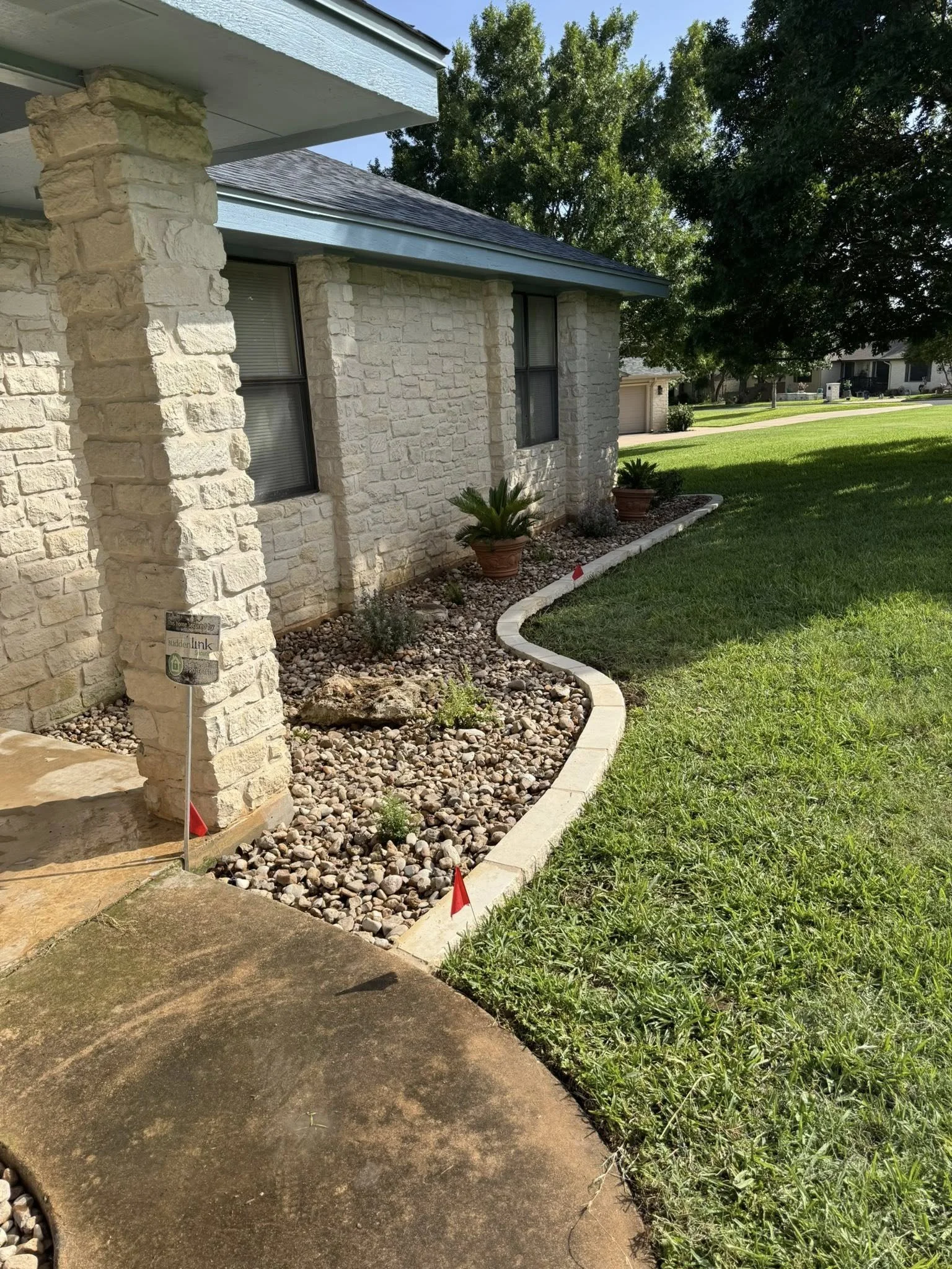 A garden bed with rocks and plants bordered by concrete edging in front of a stone house with two windows and a blue trim over a lush green lawn.