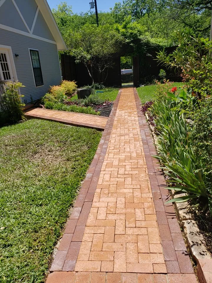 Brick walkway leading to a black gate in a backyard, surrounded by green grass, plants, and trees.