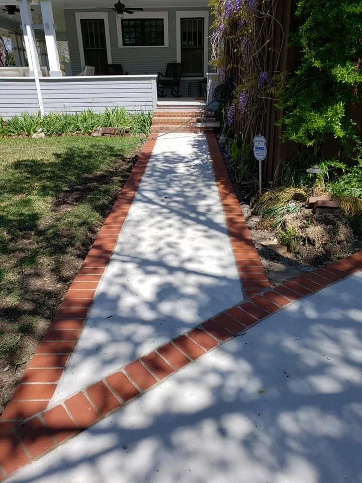 Concrete sidewalk with red brick border leading to porch of a house, with plants and greenery on the sides.