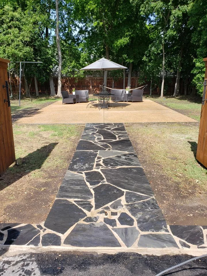 Backyard patio with a stone path leading to a seating area under an umbrella, surrounded by trees and a wooden fence.