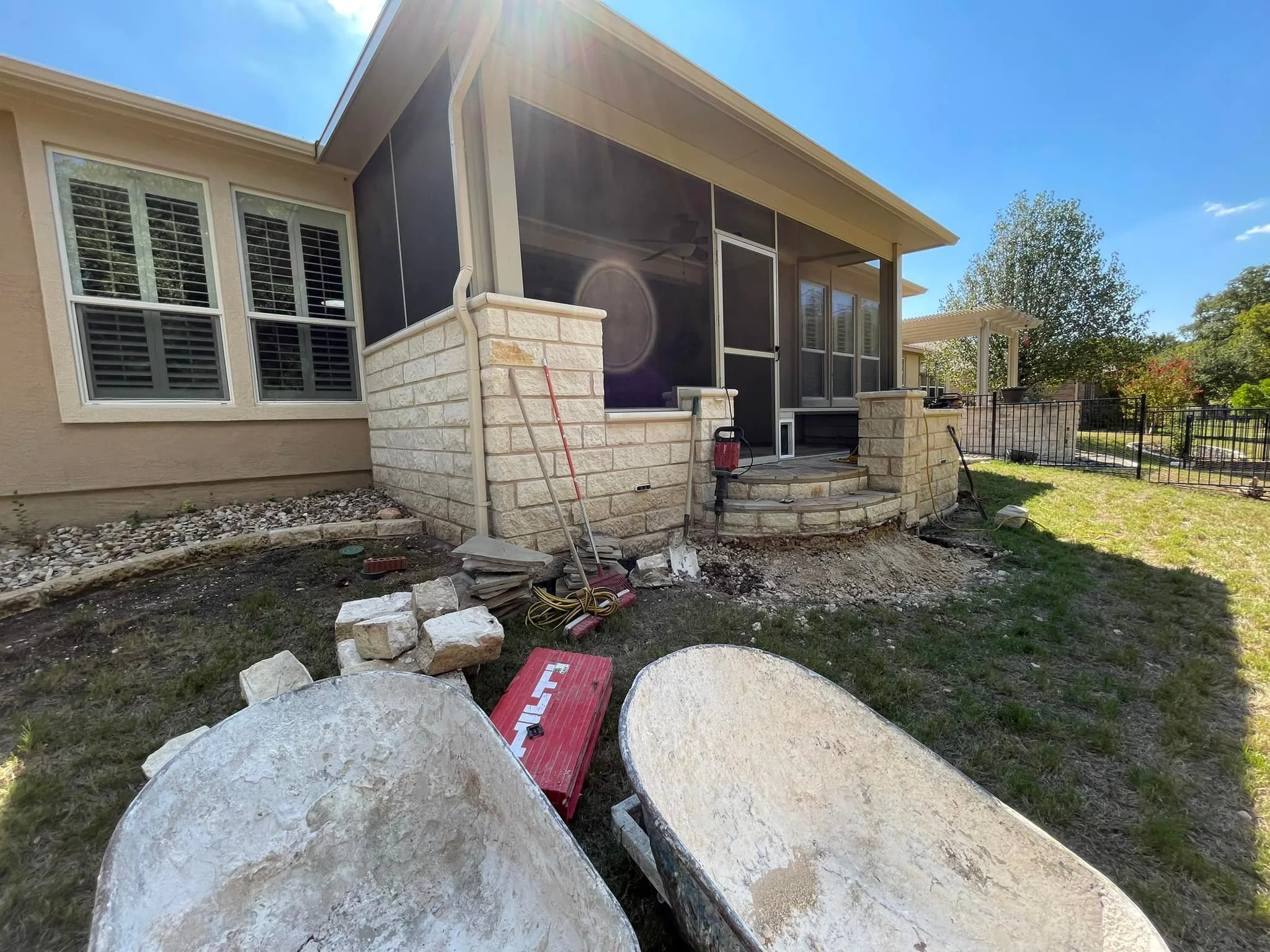 Backyard patio under renovation with construction tools, stools, and dirt, showing a screened porch, a stone patio, and a grassy yard with trees in the background.