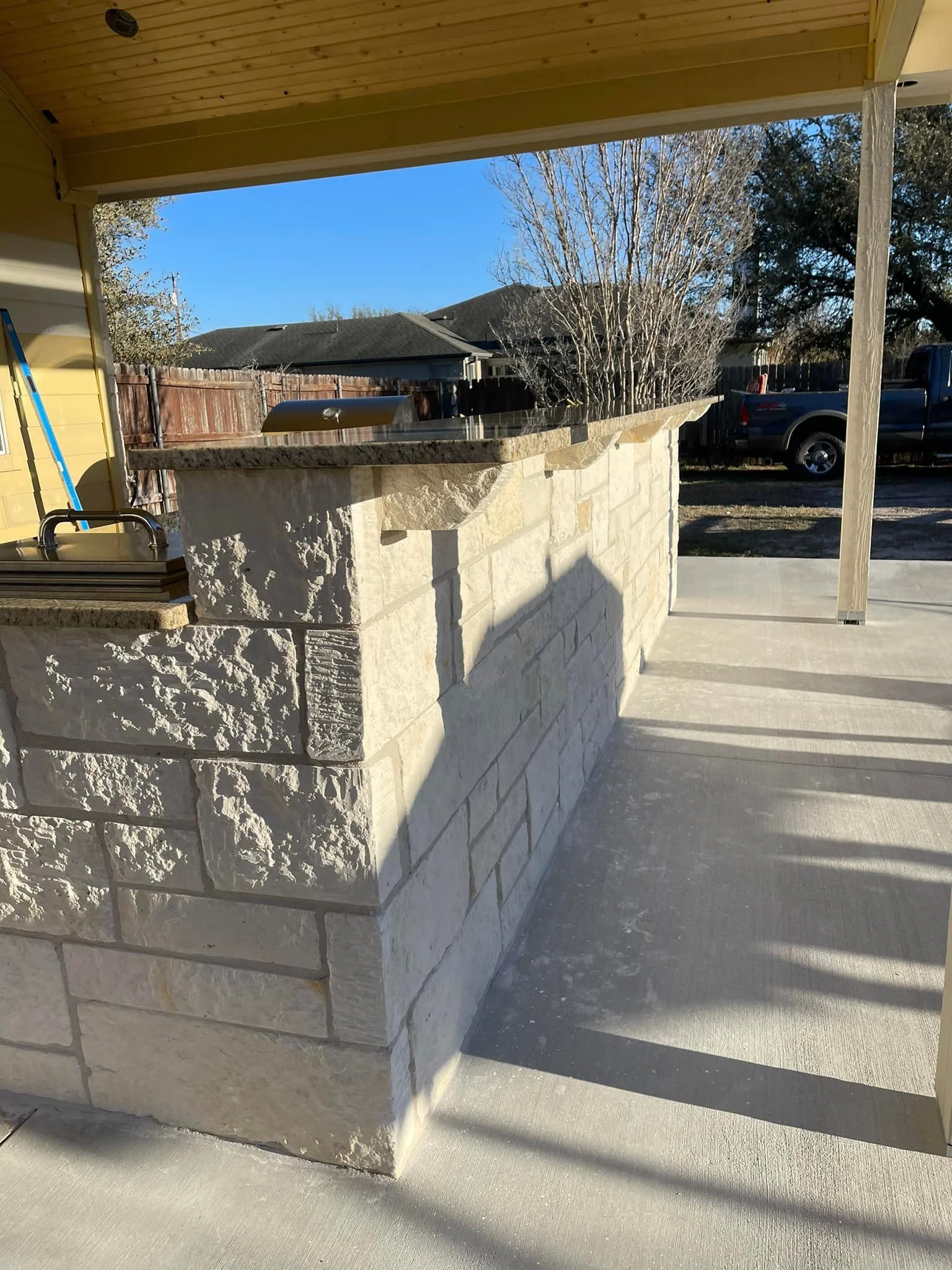 An outdoor kitchen counter with a stone base and granite countertop, with a sink and a barbecue grill, under a covered patio near a backyard with a wooden fence, trees, and a parked vehicle.