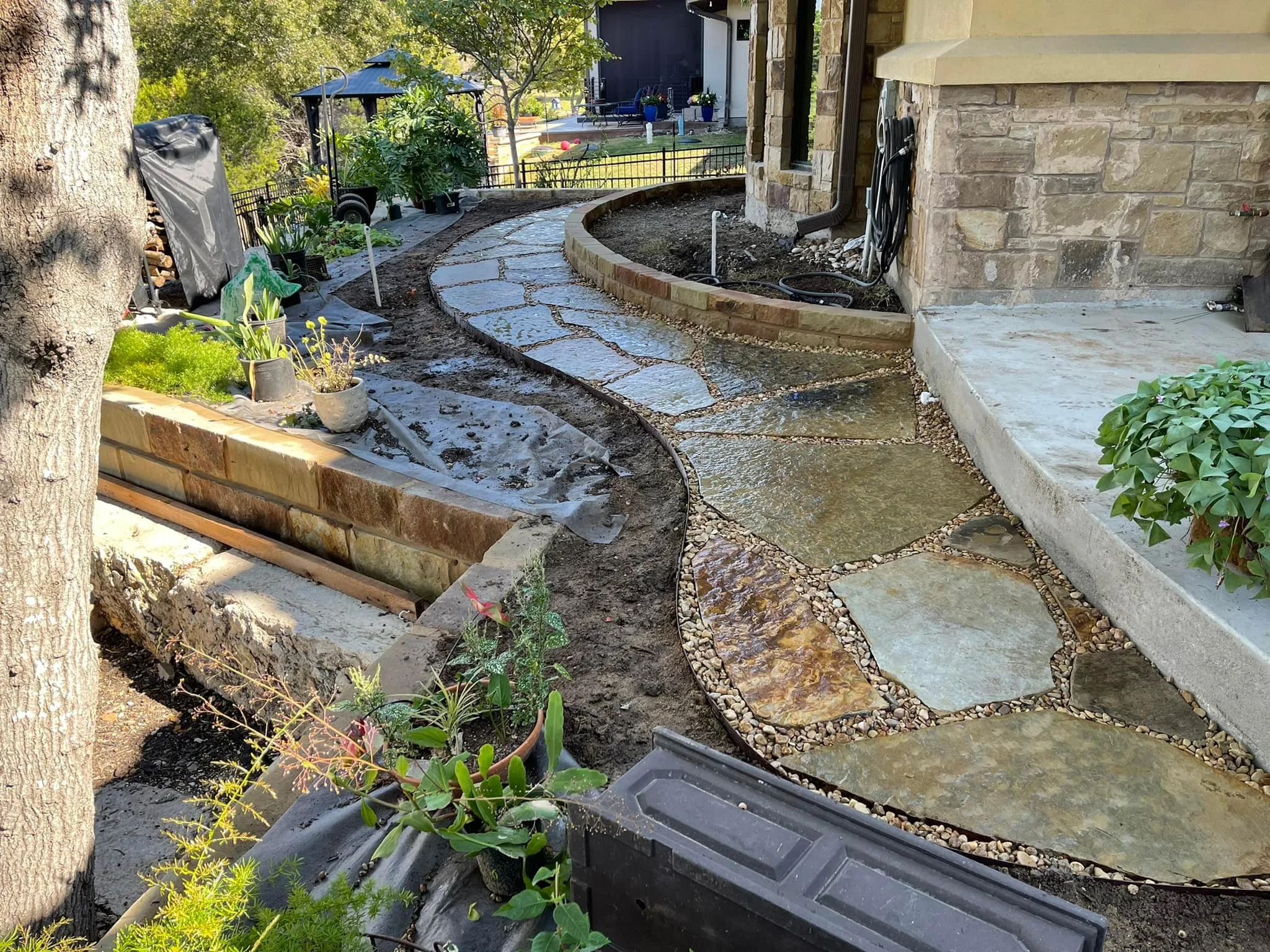 Curving stone pathway being installed next to a house, with ongoing landscape work and various potted plants nearby, in a backyard.