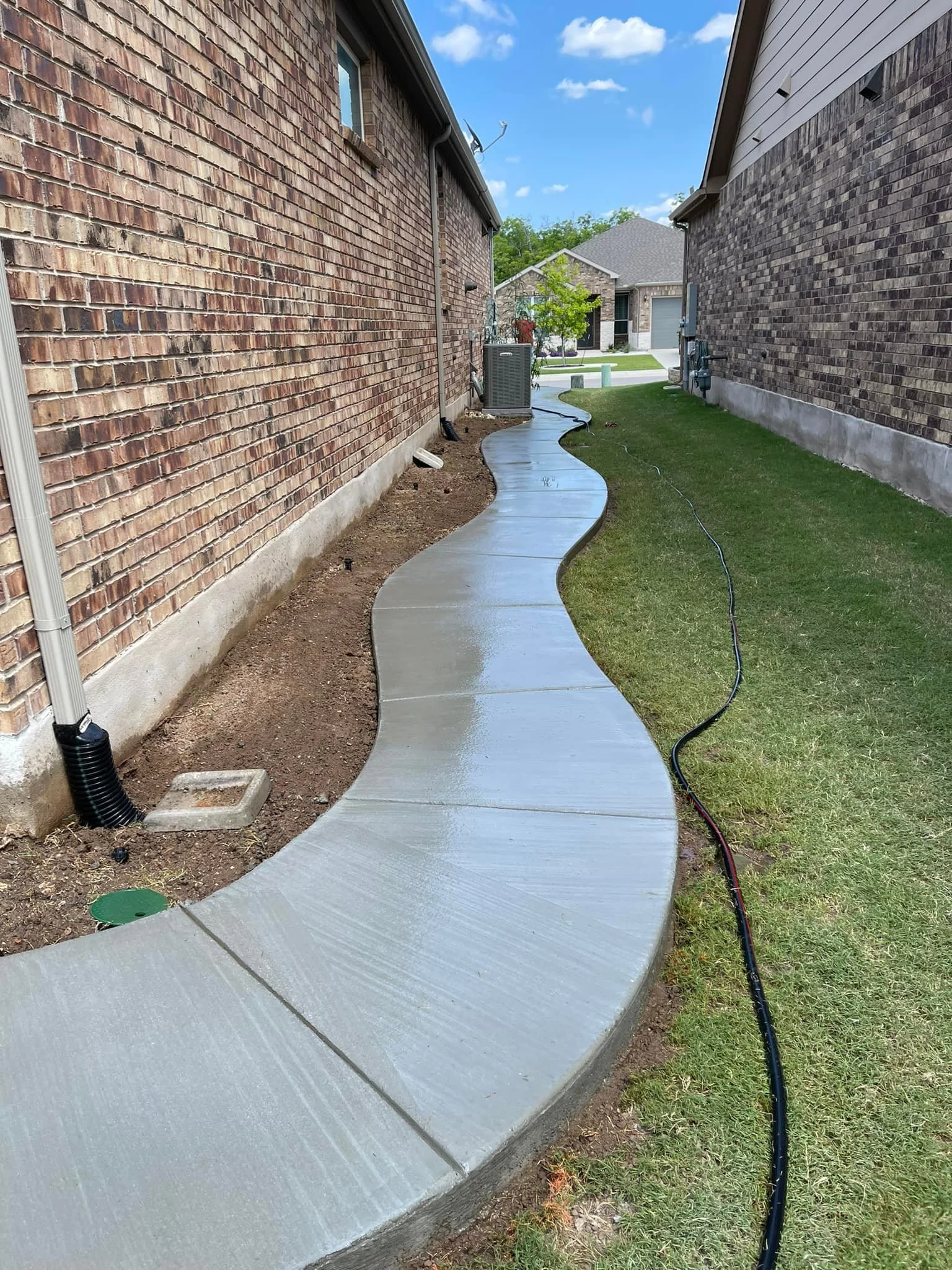 Recently poured concrete sidewalk with a wavy design between two brick houses, lawn on one side, construction tools and hoses nearby, under a blue sky with some clouds.