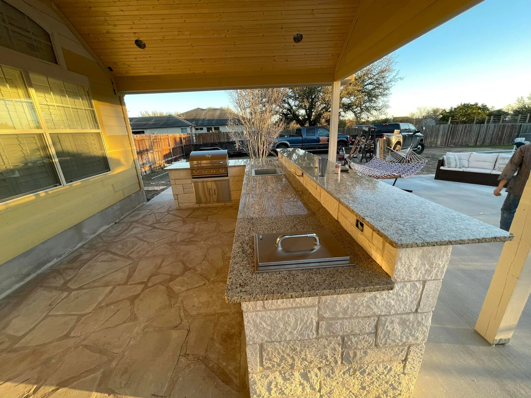 Outdoor patio with stone flooring, a granite countertop, grill, sink, and seating area with a hammock and couches, surrounded by a wooden fence and trees.