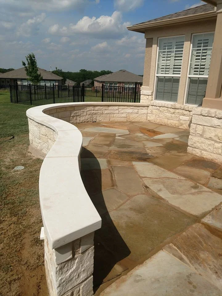 A backyard patio with a curved stone wall and a concrete bench, overlooking neighboring houses and a grassy area under a partly cloudy sky.