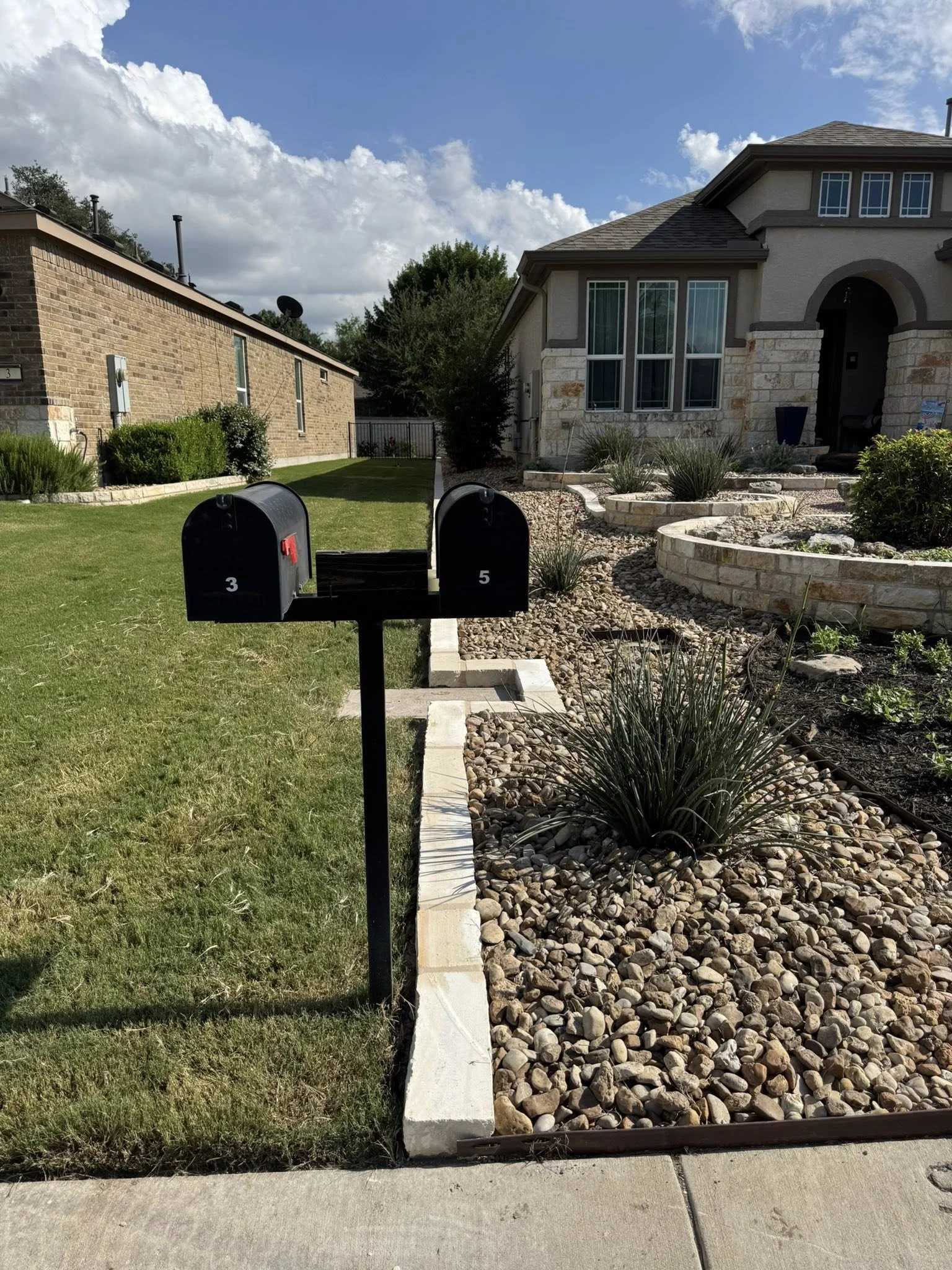 Two black mailboxes labeled 3 and 5 in front of a house with a landscaped yard with rocks, grass, and plants, under a partly cloudy sky.
