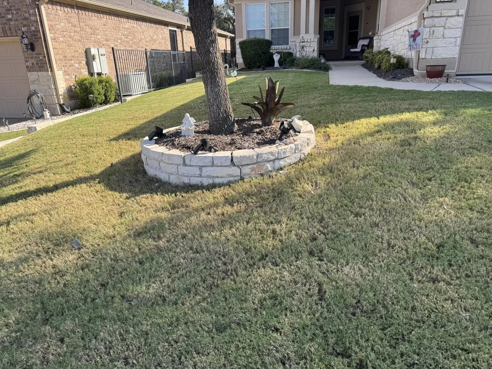A front yard with a circular stone garden bed containing a tree and a plant, decorated with garden lights and garden ornaments. A house with a porch is in the background.