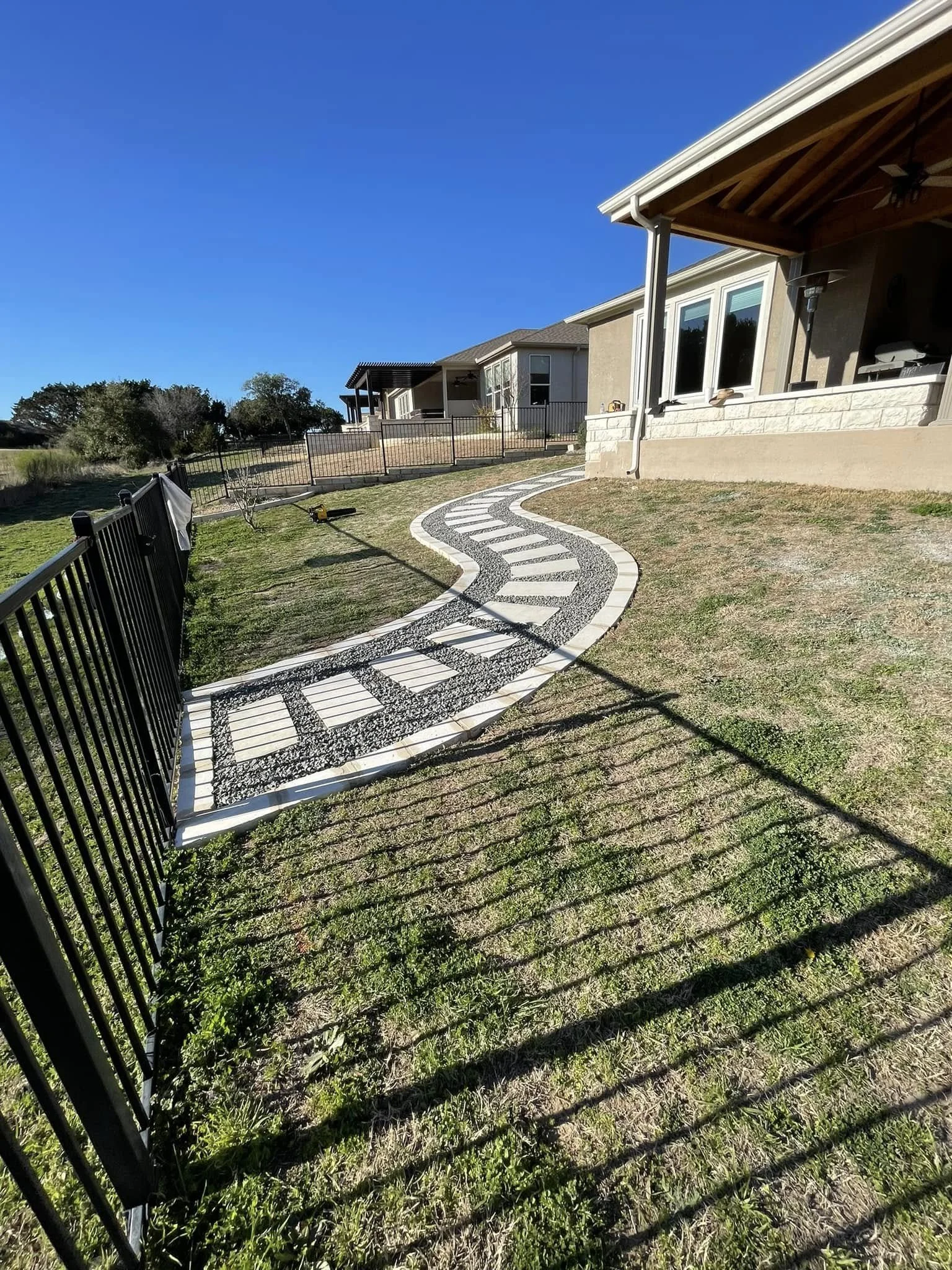 A curved pathway with white bricks and dark gravel leading from a backyard gate toward a house with a porch, under a clear blue sky.