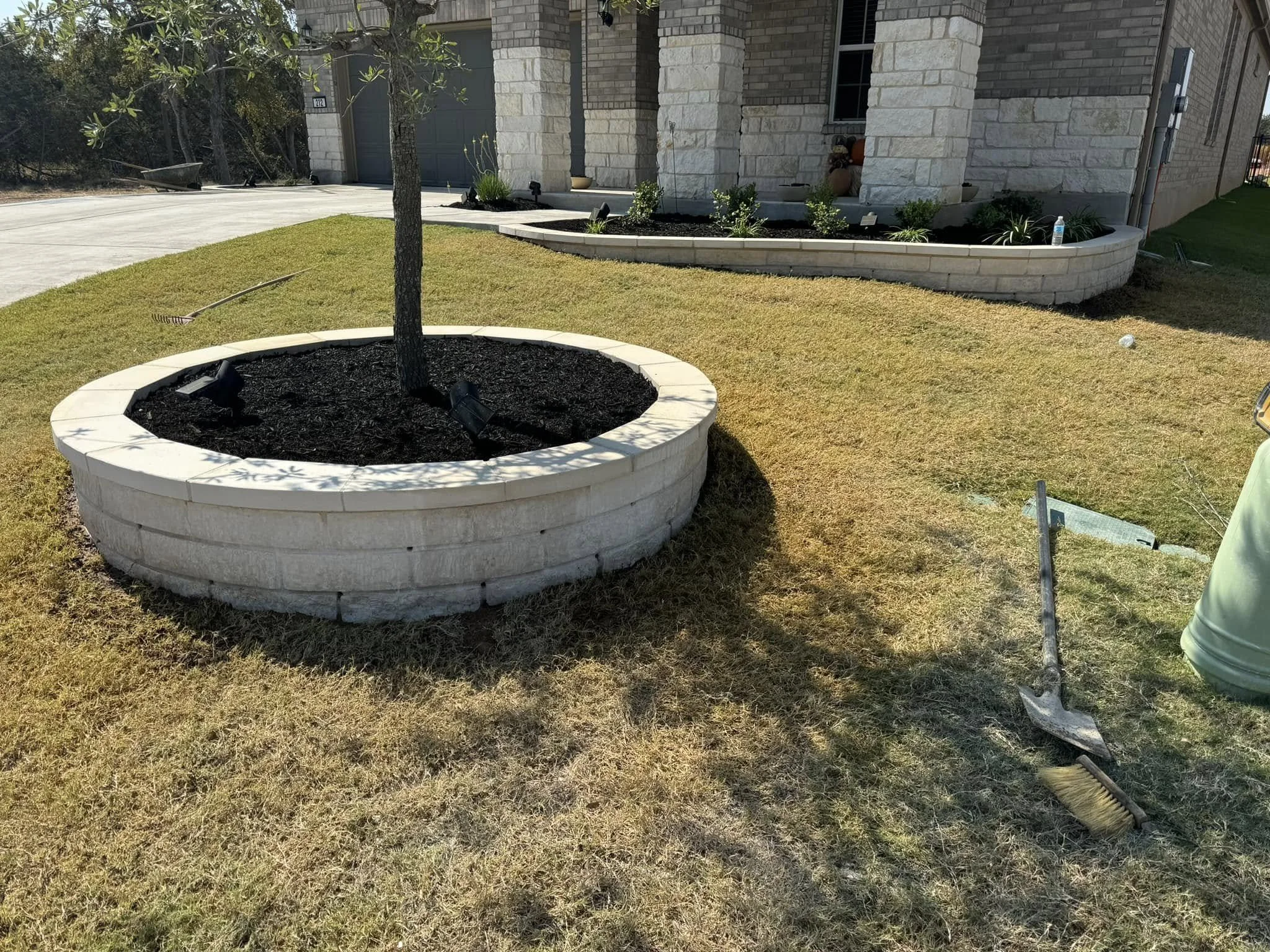 A landscaped front yard of a house with a small tree in a round stone planter, freshly planted with dark soil. A rake and a broom lie on the grass, as well as a green trash bin nearby. The house has stone exterior walls and a small garden bed with pl