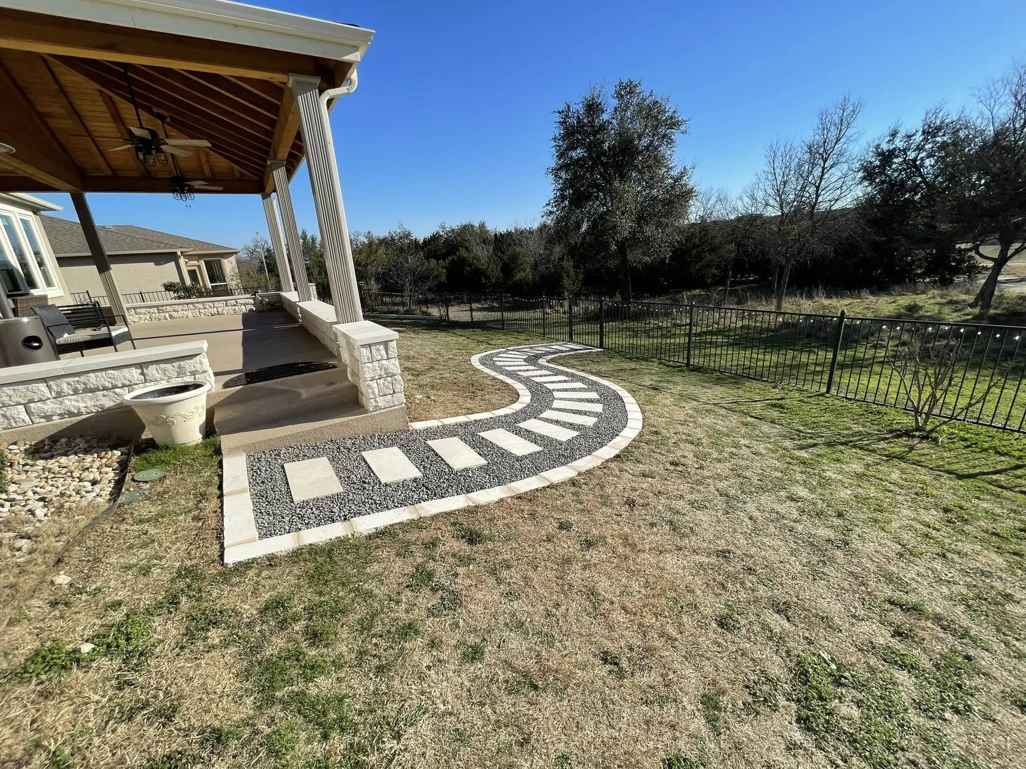 Backyard with a paved pathway, grassy lawn, porch with columns, and trees in the background.
