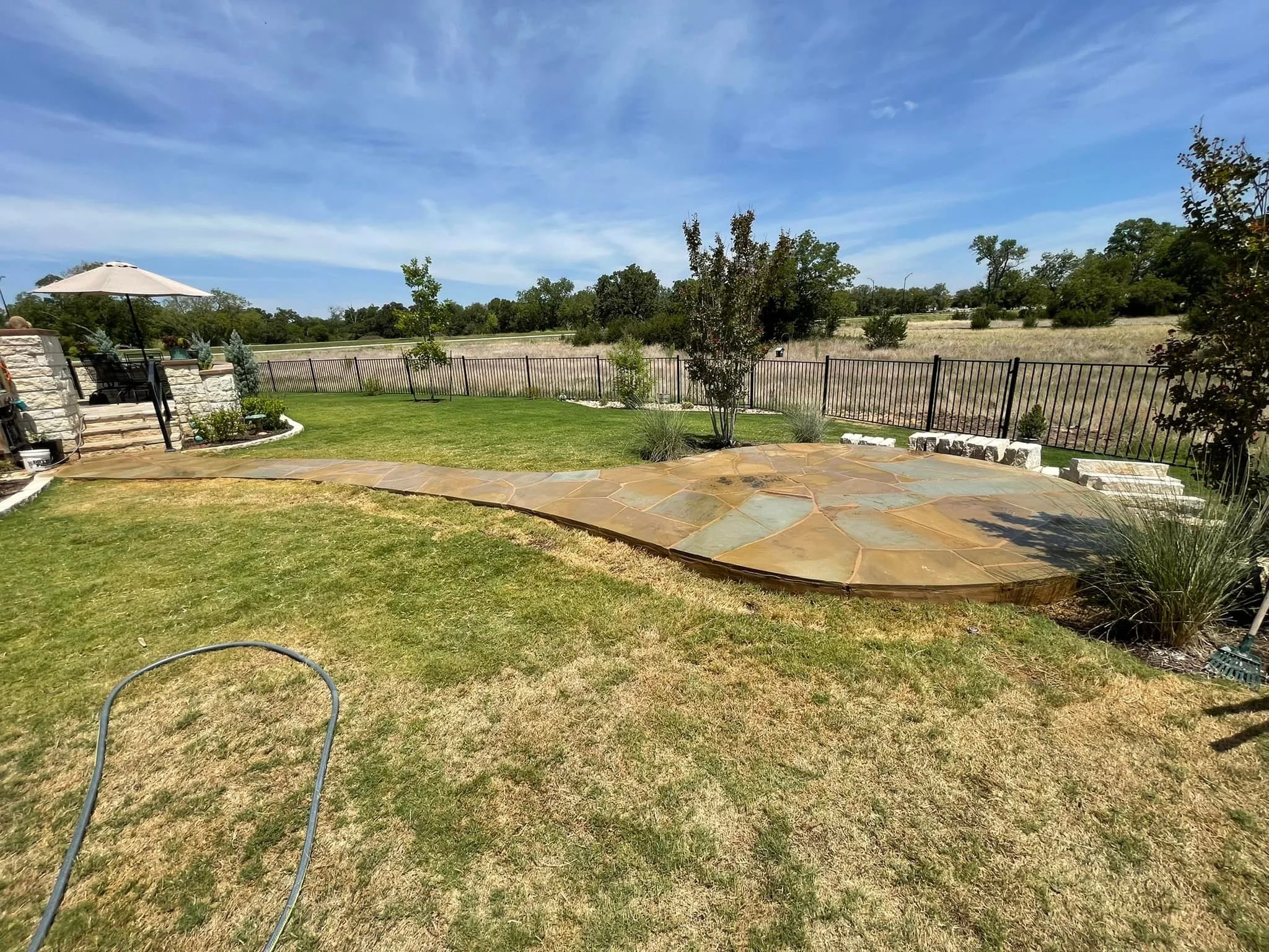 Backyard with a curved, multi-colored stone patio, green grass, small trees, decorative bushes, a black metal fence, and an open field beyond under a partly cloudy sky.