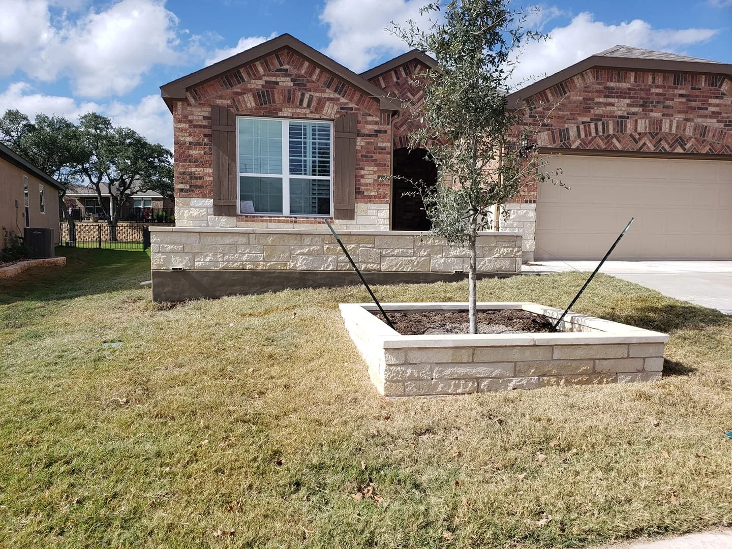 A brick house with a small front yard features a young tree planted in a white-painted brick planter, with two black support stakes attached to the tree, and a beige garage door on the right side. The sky is partly cloudy.