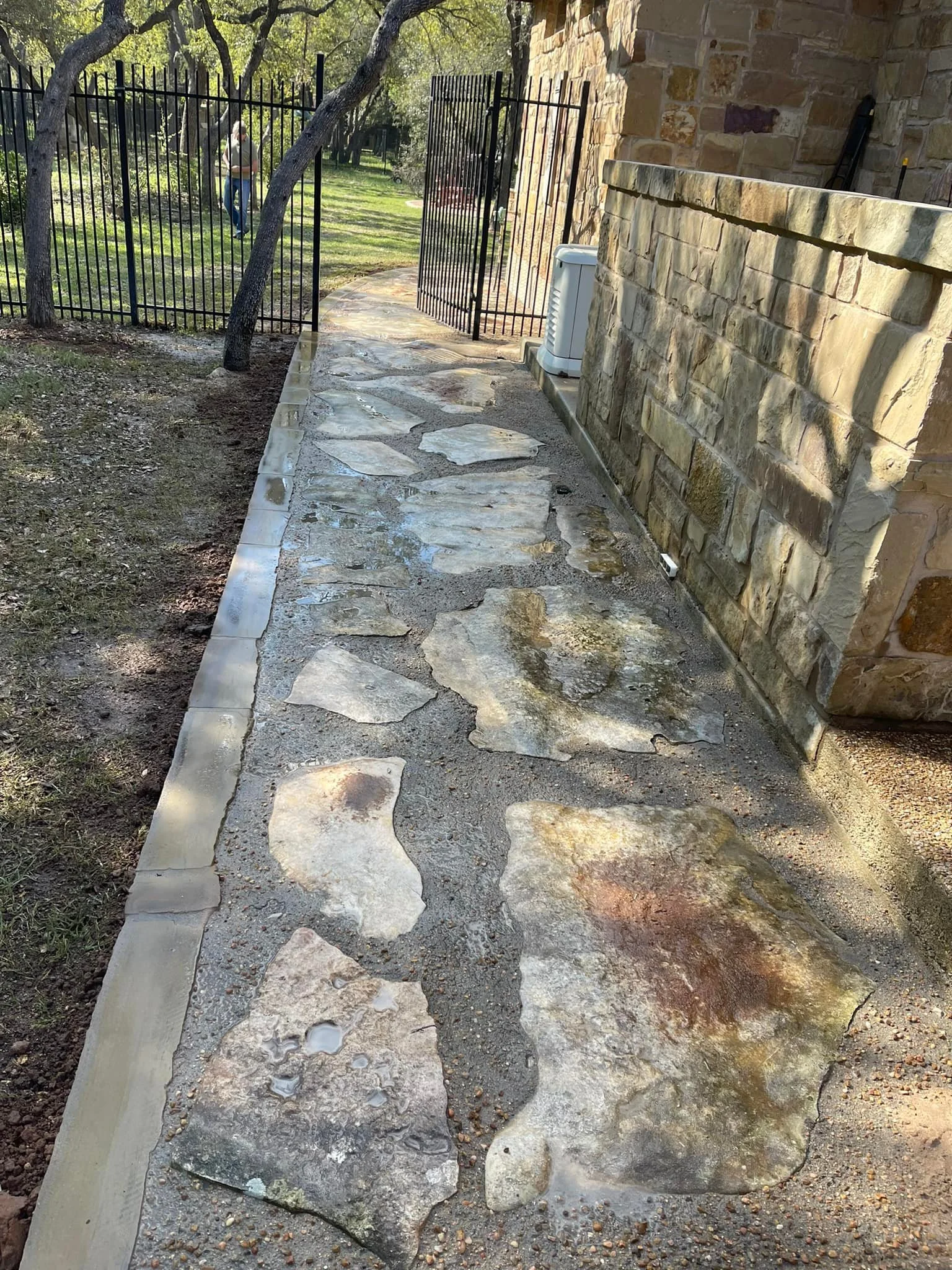 A stone pathway runs alongside a house with a stone exterior wall. To the left, there is a grassy yard with trees and a person walking in the background. The pathway is bordered by a curb and is wet.