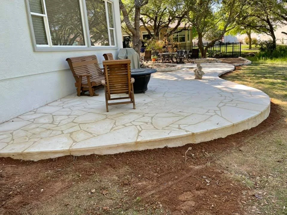 A backyard patio area with a curved, light-colored stone surface, surrounded by grass and trees, with outdoor furniture including chairs, a table, and a barbecue grill.