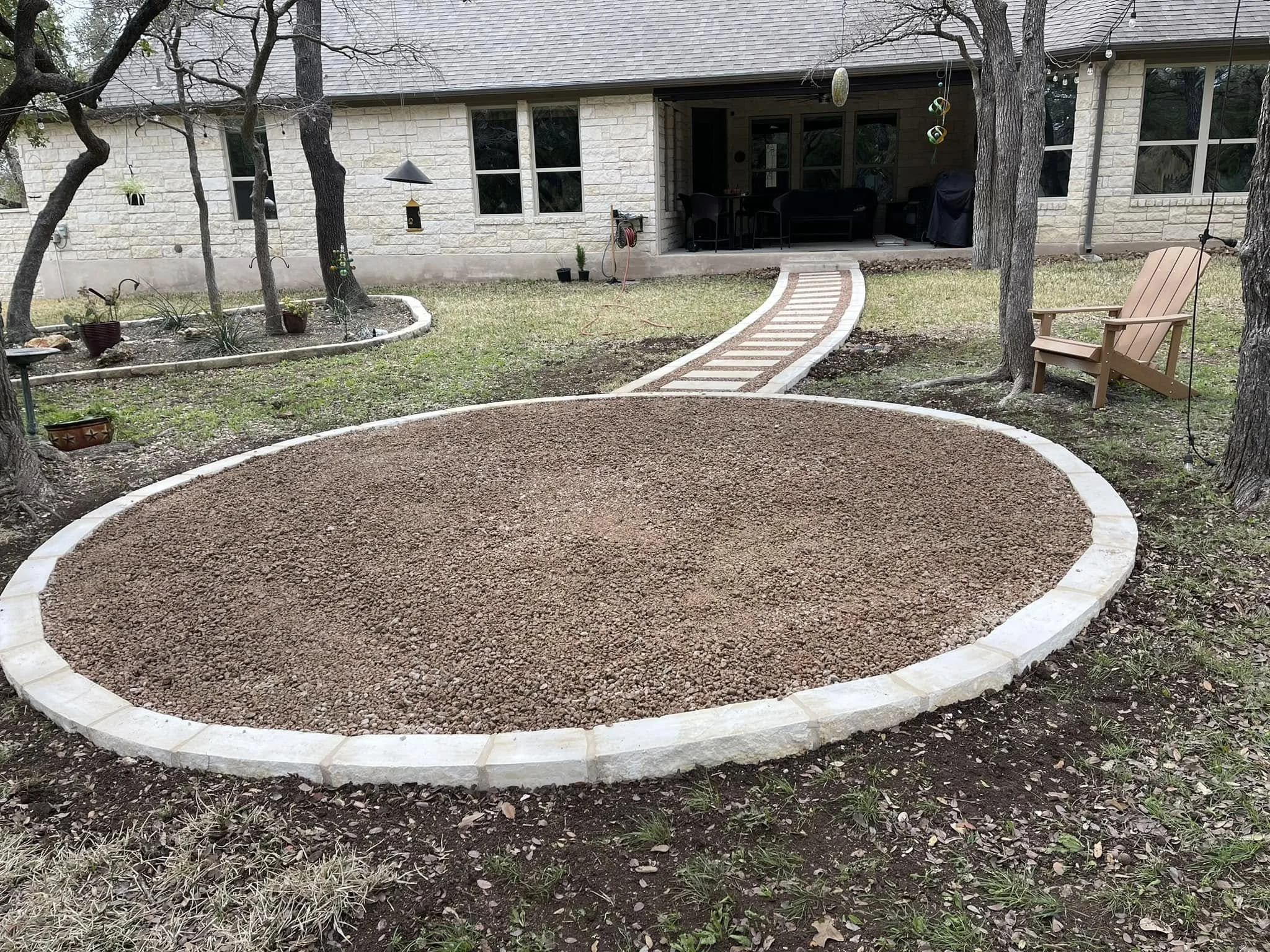 A backyard with a curved brick border surrounding a gravel area, with a wooden walkway leading from the house to the yard. There are trees, potted plants, a wooden chair, and a covered patio in the background.