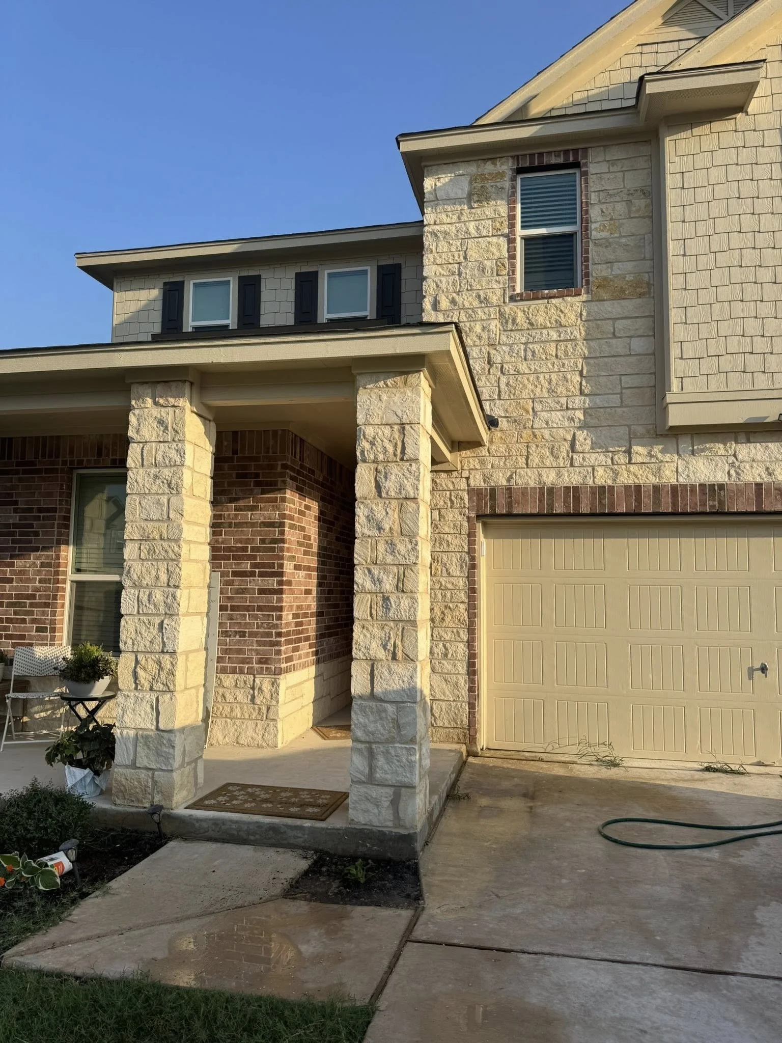Front view of a house with a brick and stone facade, a porch supported by stone columns, and a yellow garage door. There is a garden hose on the driveway and a small potted plant on a chair near the house.