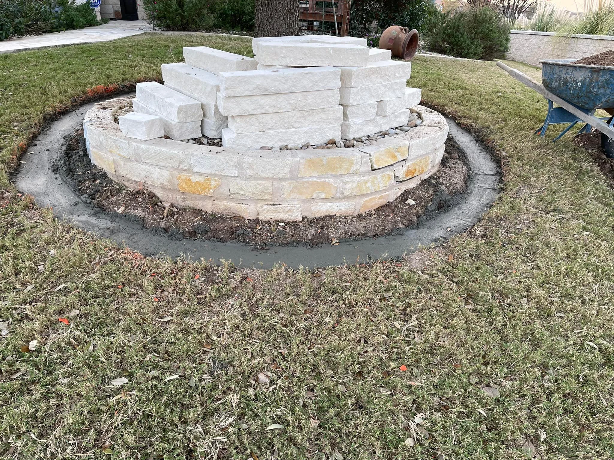 A circular brick and stone fire pit under construction with stacked bricks and stones, surrounded by grass and construction tools like a wheelbarrow and a clay pot.