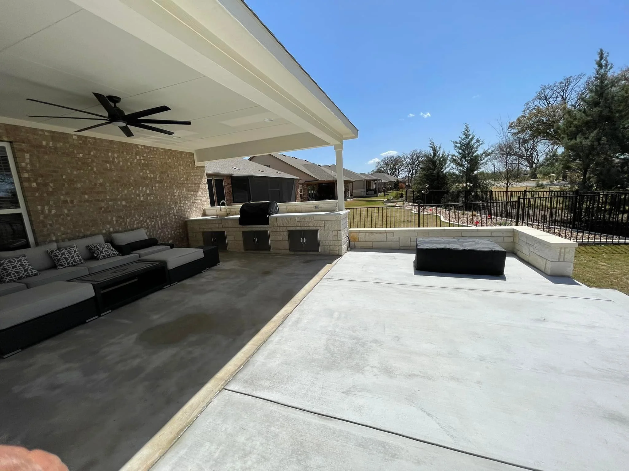 A spacious backyard patio with outdoor seating, a ceiling fan, a built-in grill, and a view of the backyard lawn and trees under a blue sky.