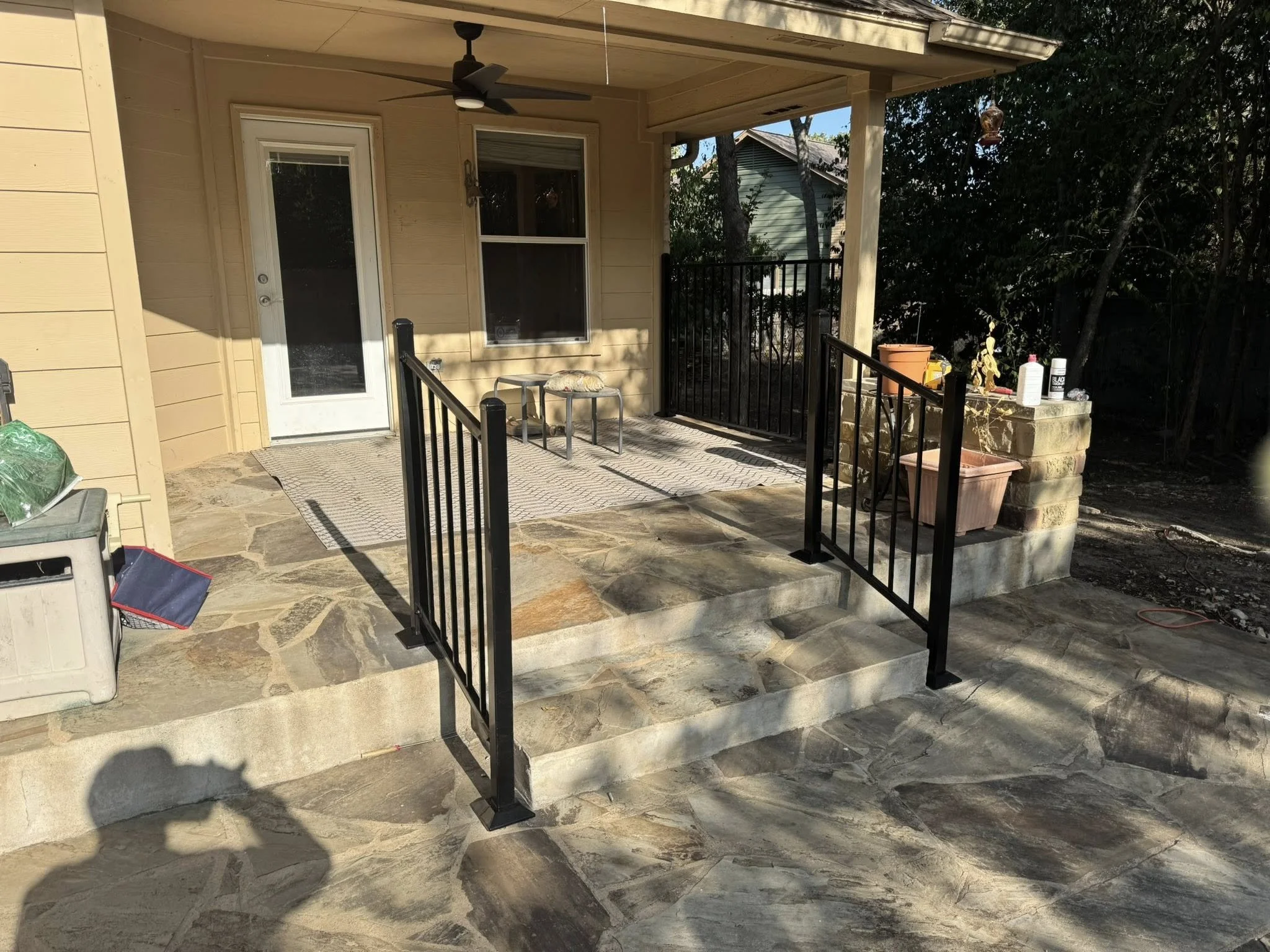 View of a small porch with a stone floor, black metal railing, and a yellow house exterior. The porch has a ceiling fan and a door leading inside. There are some garden tools and plants on the right side, and a small bench with a pillow on it in fron