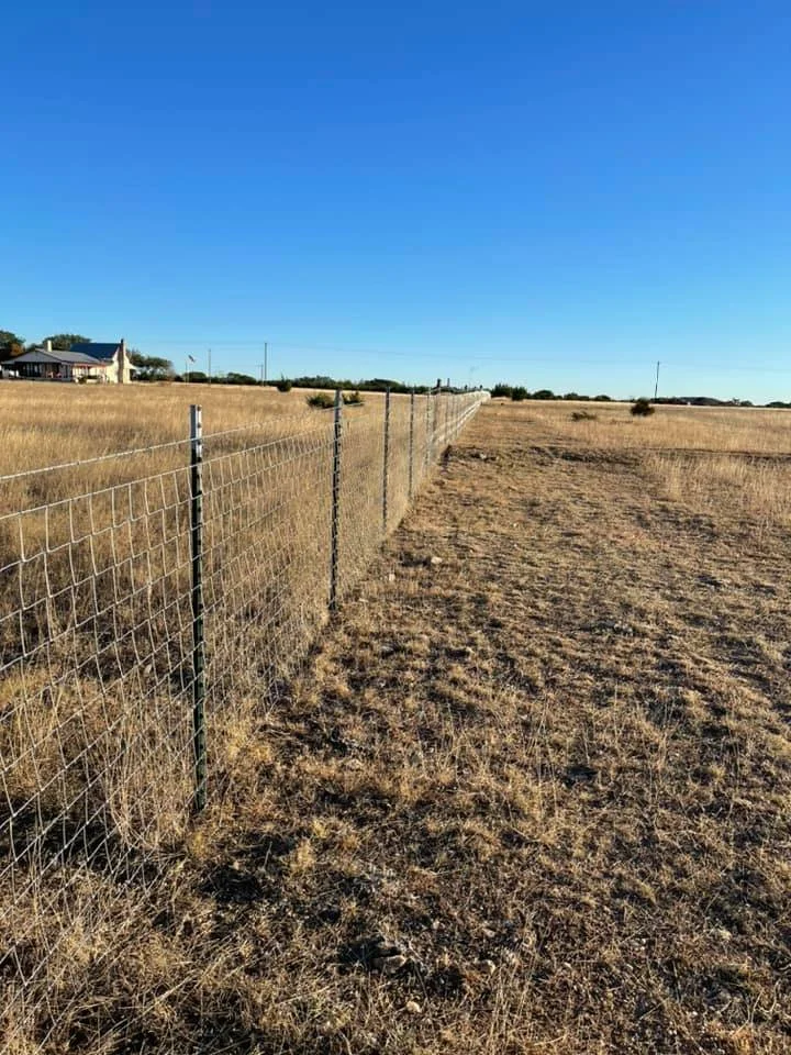 A dirt field with dry grass and weeds, enclosed by a chain-link fence that extends towards the horizon under a clear blue sky. There is a house and some power lines in the distance.