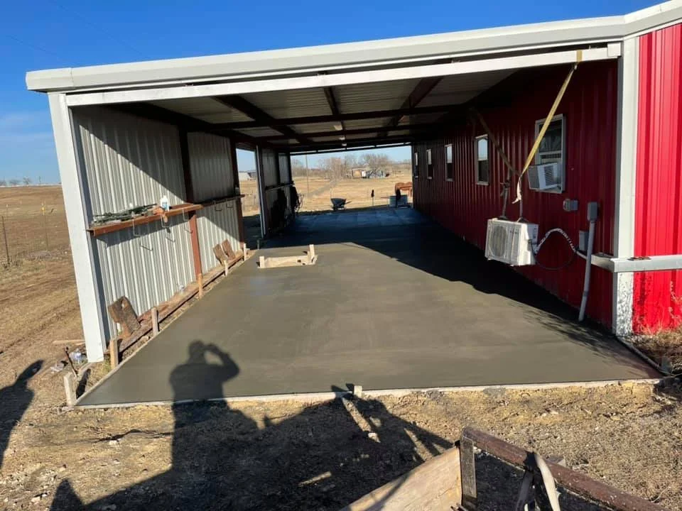 This is a partially enclosed structure with a metal roof, red side walls, and a freshly poured concrete floor. Construction tools and materials are visible, and the area appears to be in a rural setting.