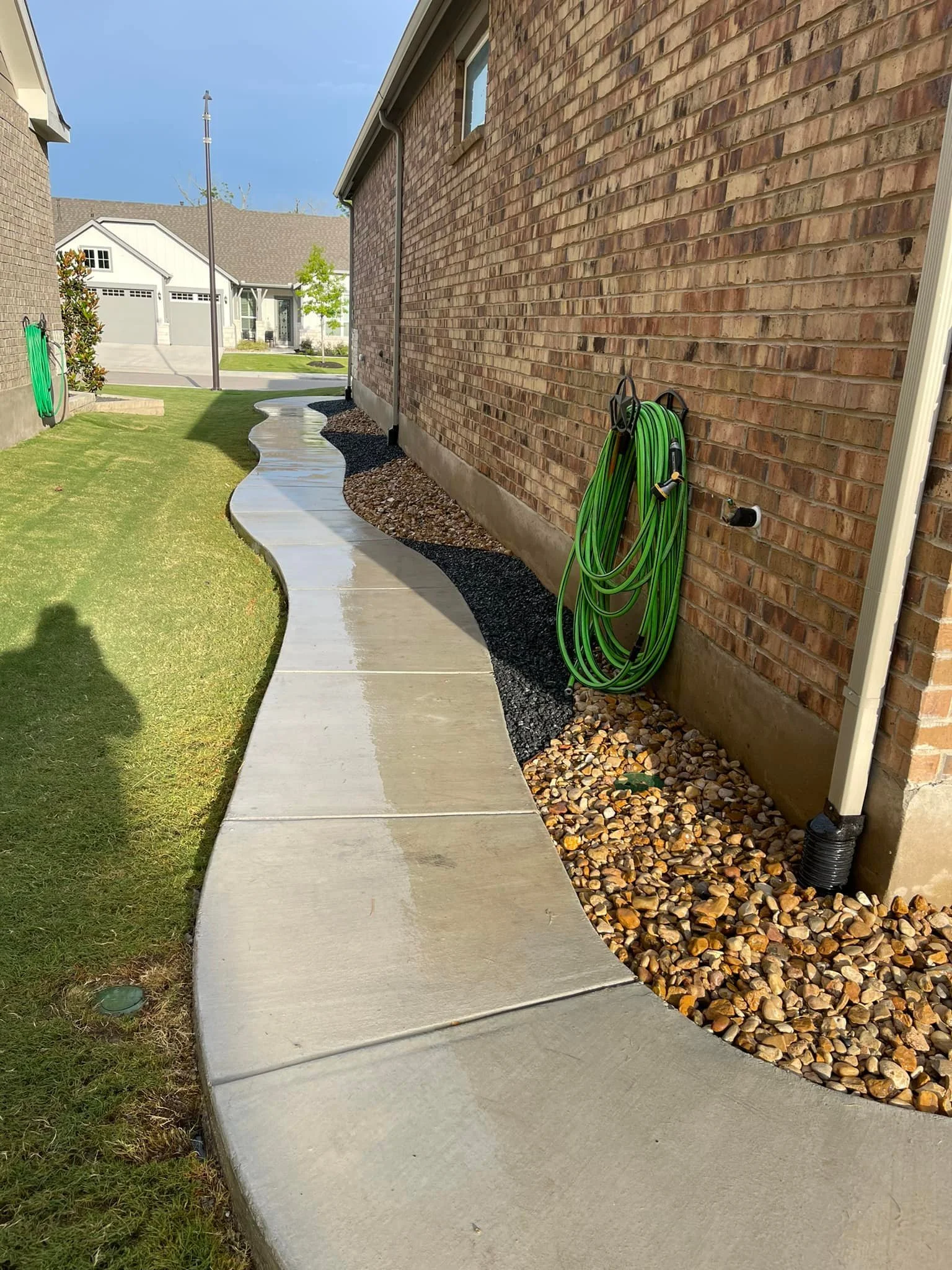 Curving concrete sidewalk running alongside a brick house, with a green garden hose hung on the exterior wall and a strip of rocks and mulch beside the house. The grass lawn is to the left.