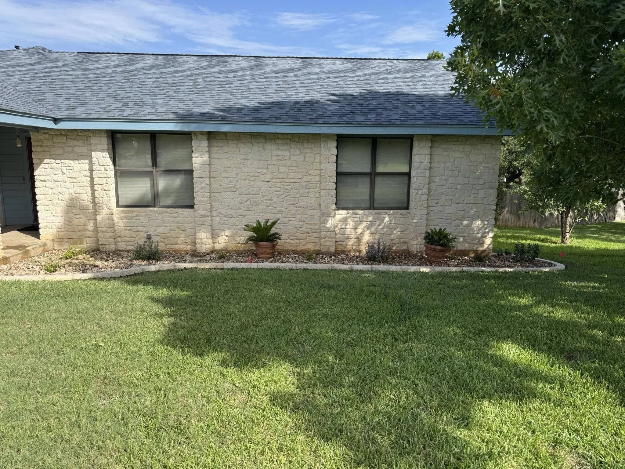 A residential house with a stone exterior, two windows, a gray shingle roof, and a well-maintained front yard with green grass, a tree, and potted plants.