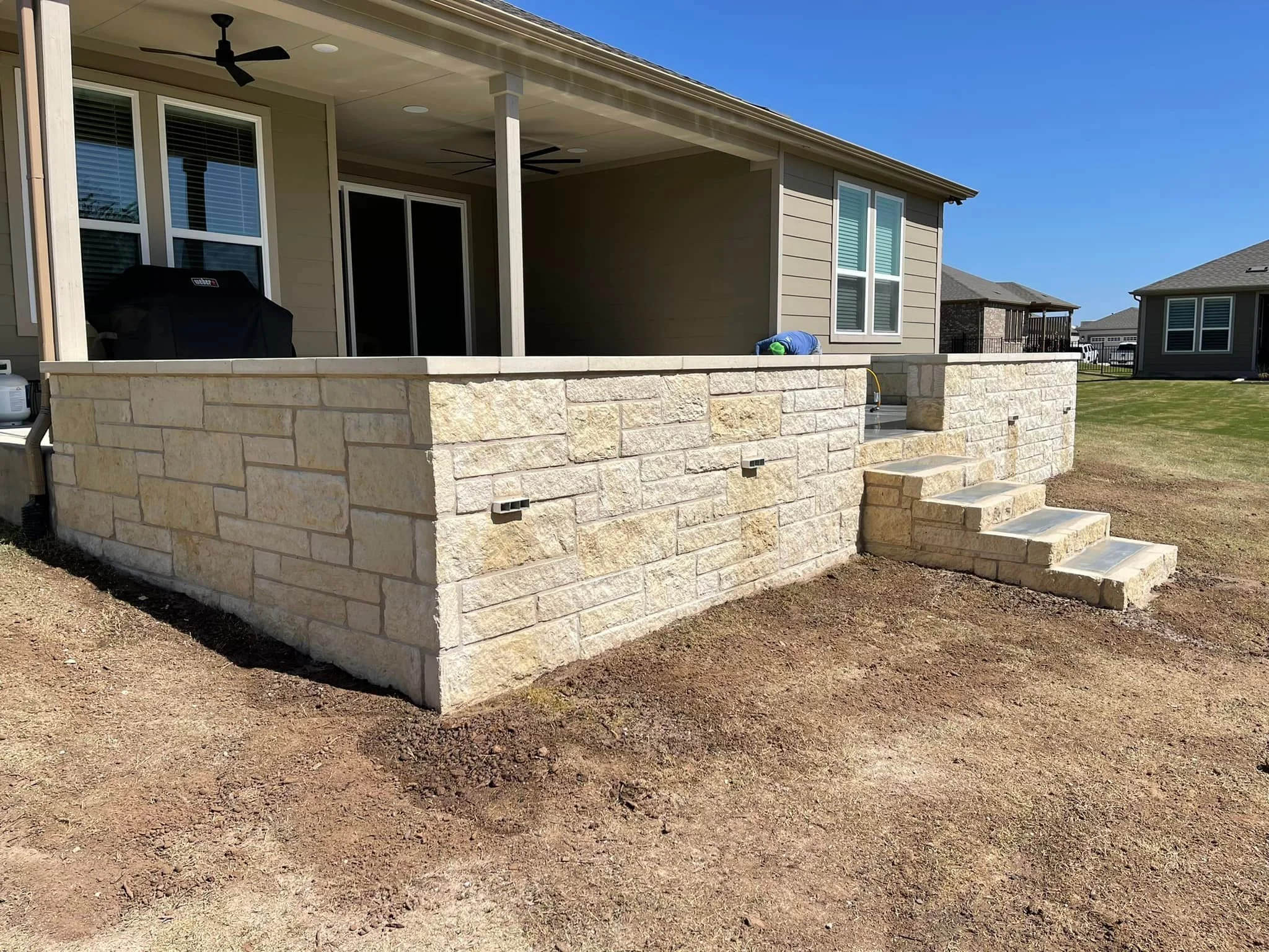 Backyard patio with stone wall, steps, and outdoor ceiling fans under a covered porch, sunny day with grass around.