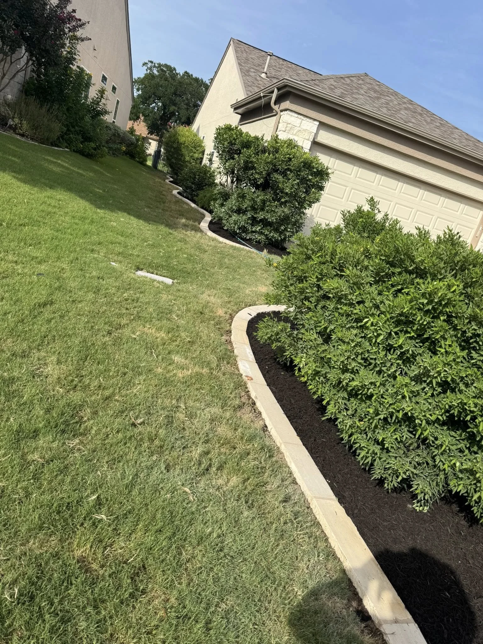 Front yard with well-maintained lawn, shrubbery, and a patio, featuring a house with a garage and a blue sky.