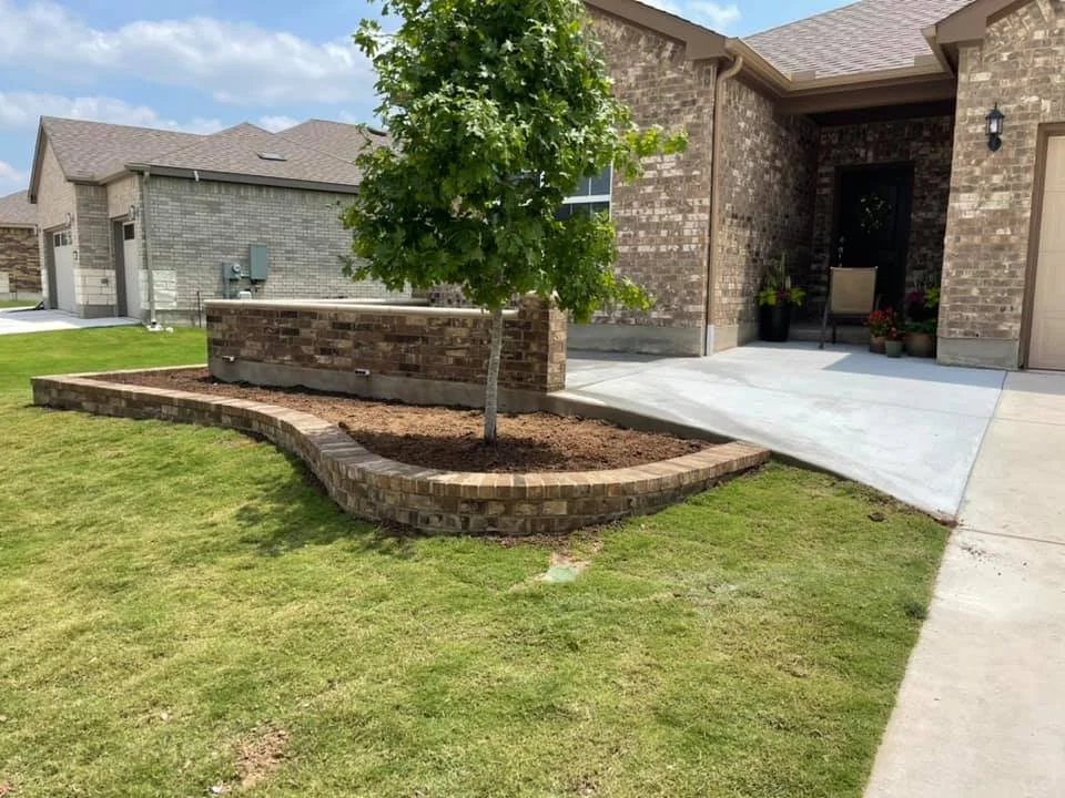 A front yard with a newly planted tree inside a brick-lined flower bed, adjacent to a concrete porch of a brick house.