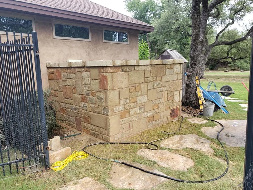 A stone wall beside a beige house with small rectangular windows, a large tree with a thick trunk, and a yard with tools and a wheelbarrow.