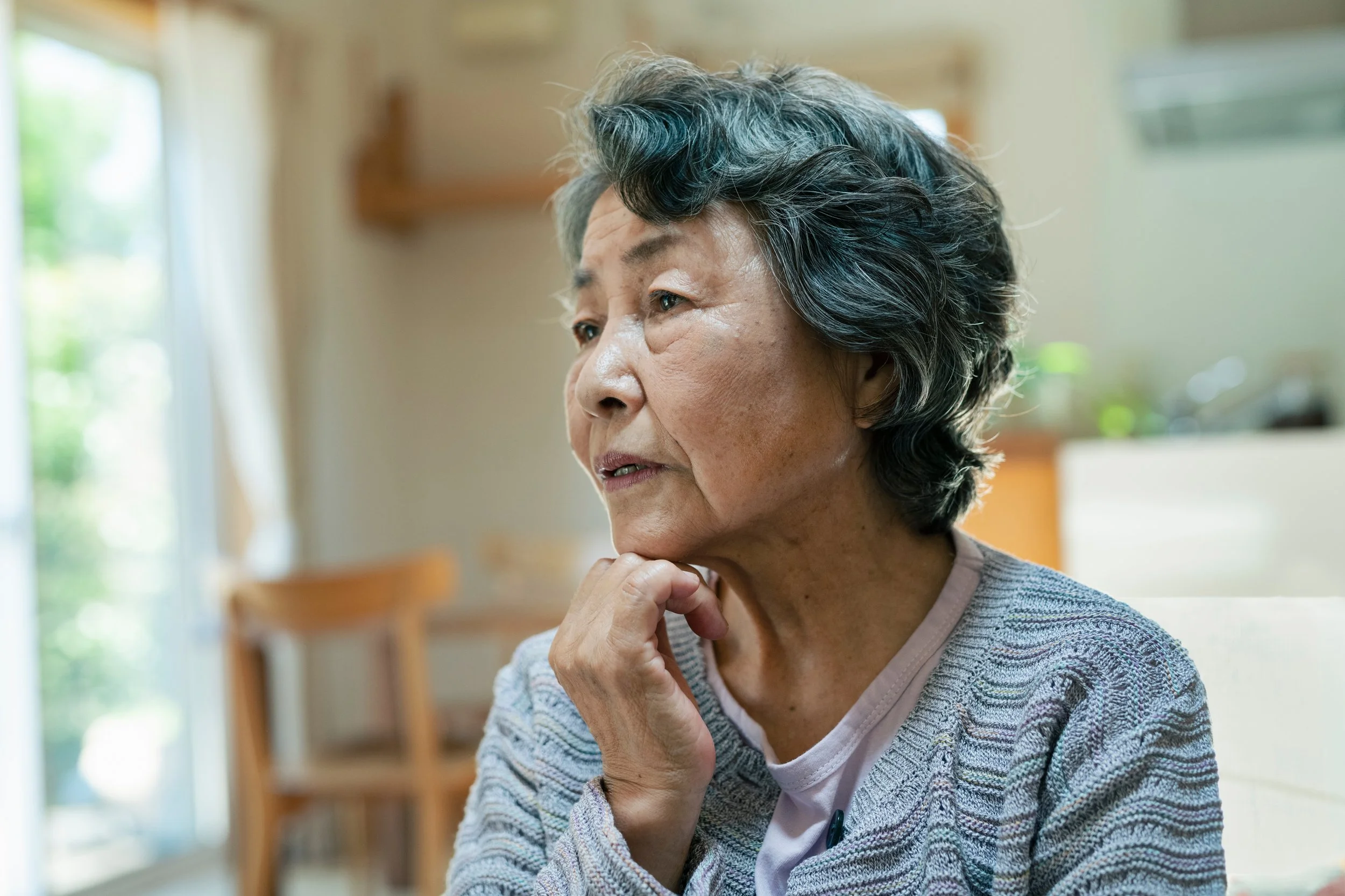 An elderly woman with gray and black hair, wearing a patterned cardigan, sitting indoors with her hand resting on her chin, gazing thoughtfully to the side.