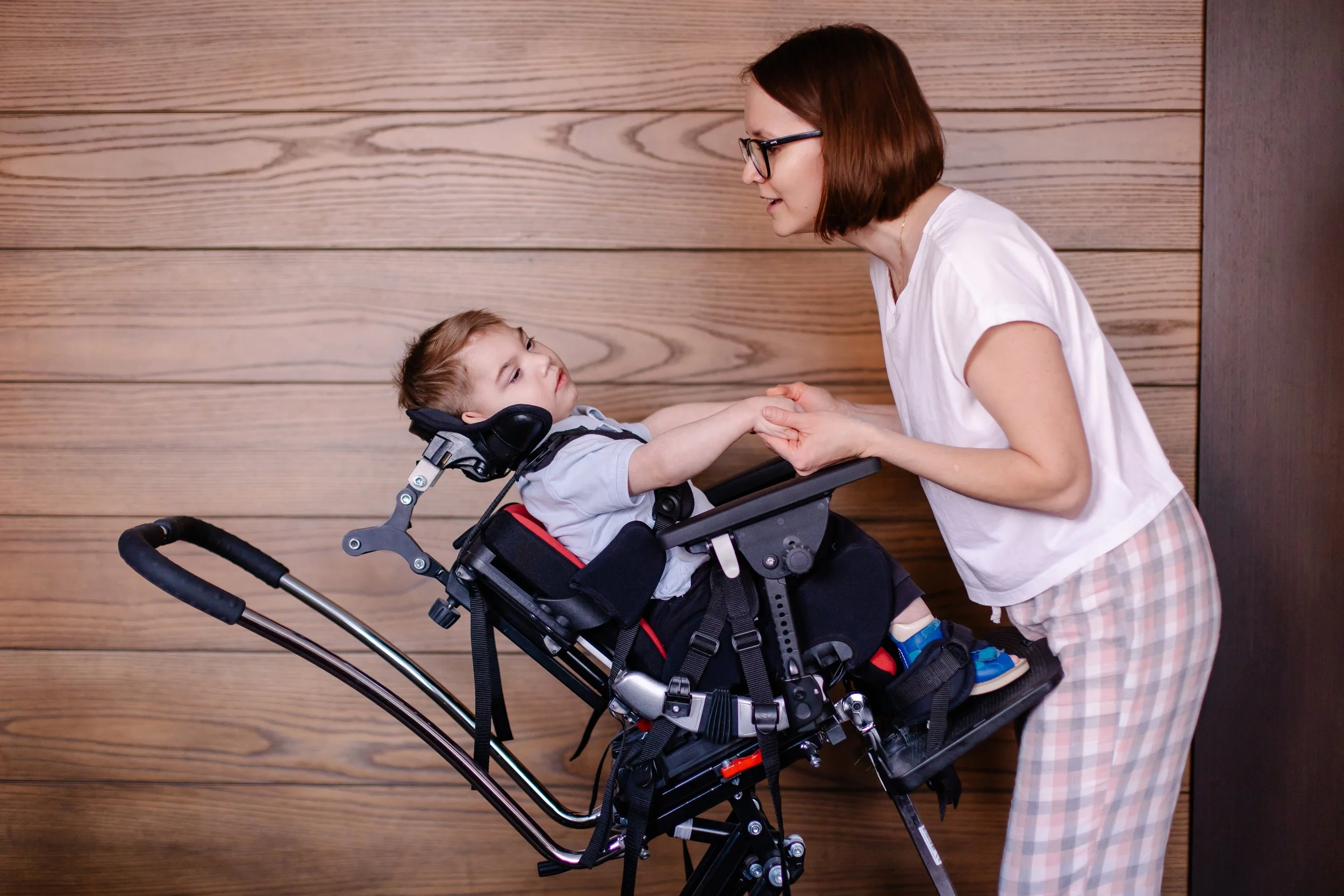 A young woman with glasses holding hands with a boy in a wheelchair against a wooden wall background.