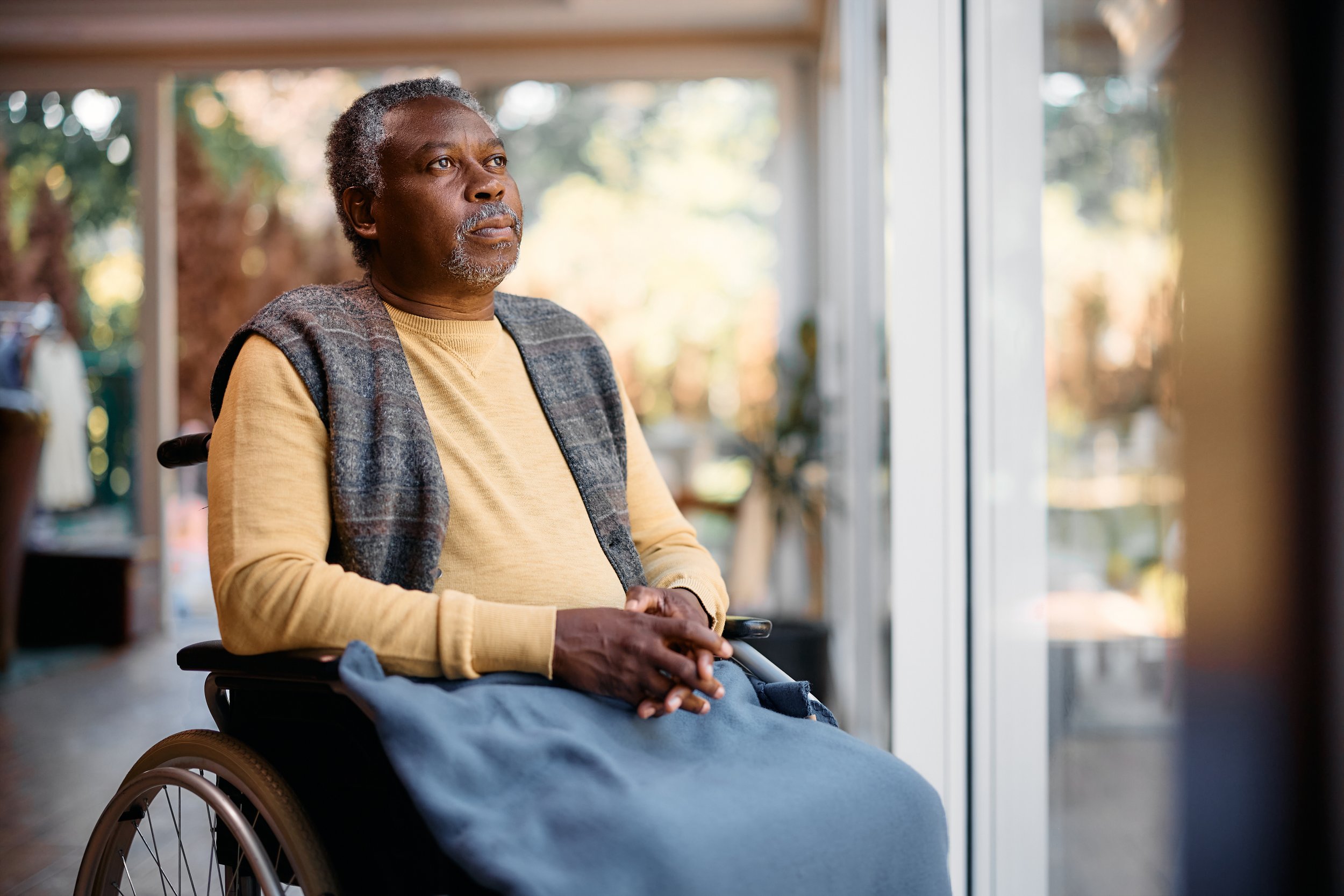 An elderly man in a wheelchair looking out a window with a thoughtful expression.