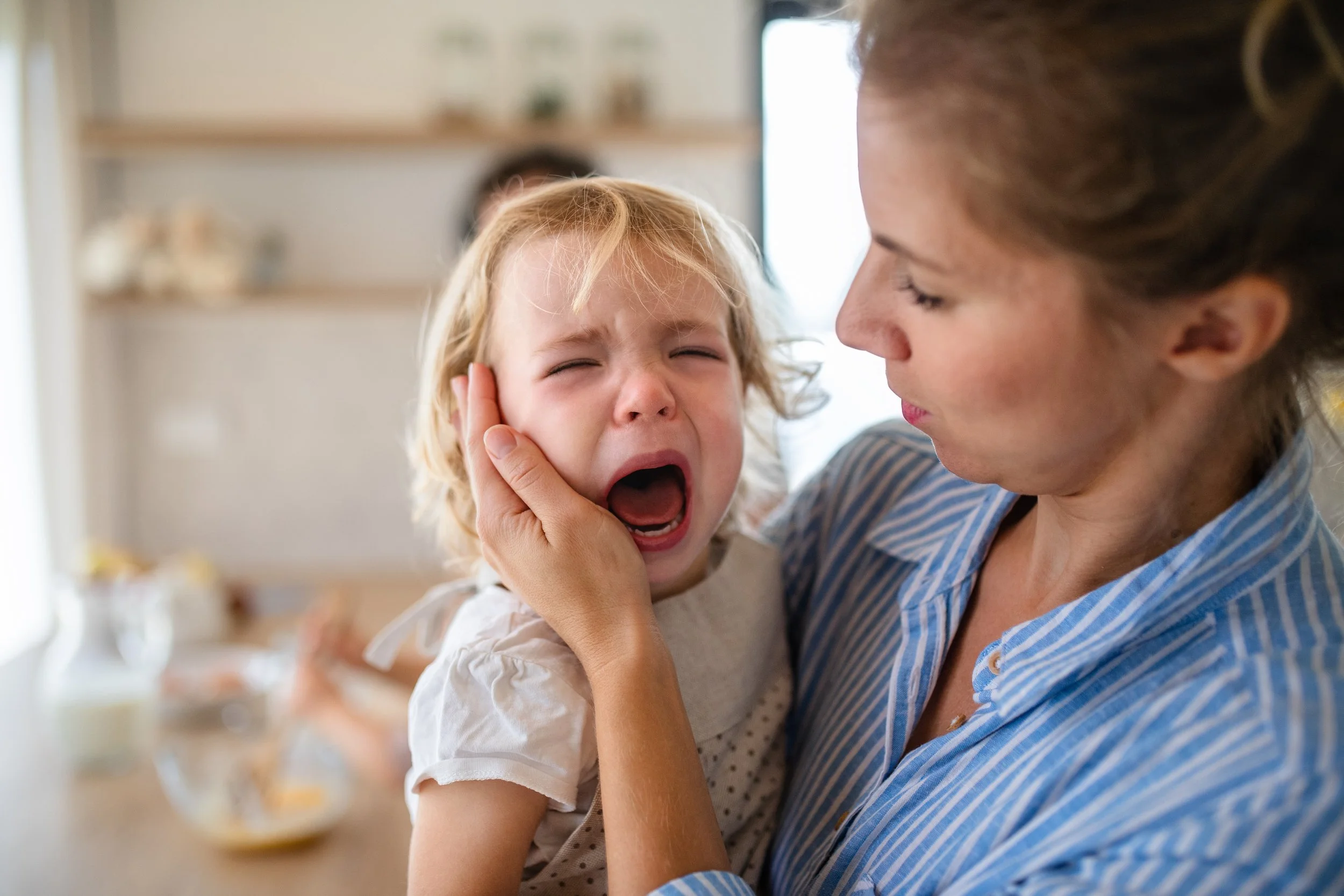 A woman holding a young girl with blonde hair, who is crying and distressed, in a bright kitchen setting.