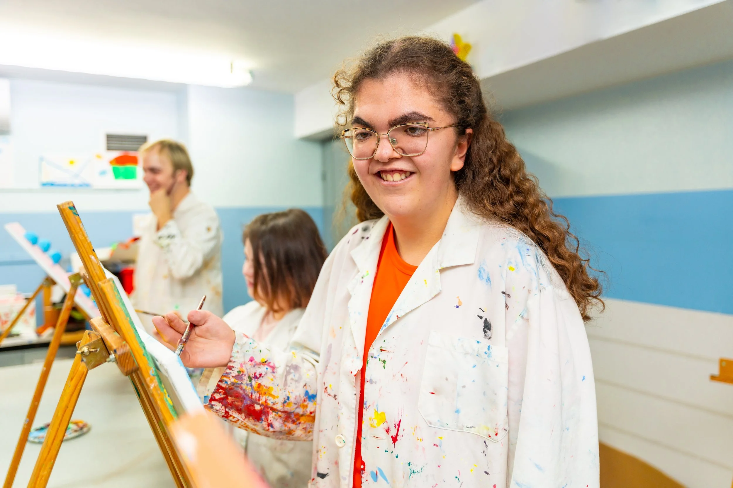 A young woman with curly brown hair, wearing glasses and a paint-splattered white apron, smiling while holding a paintbrush in her right hand, in an art classroom with other people painting in the background.