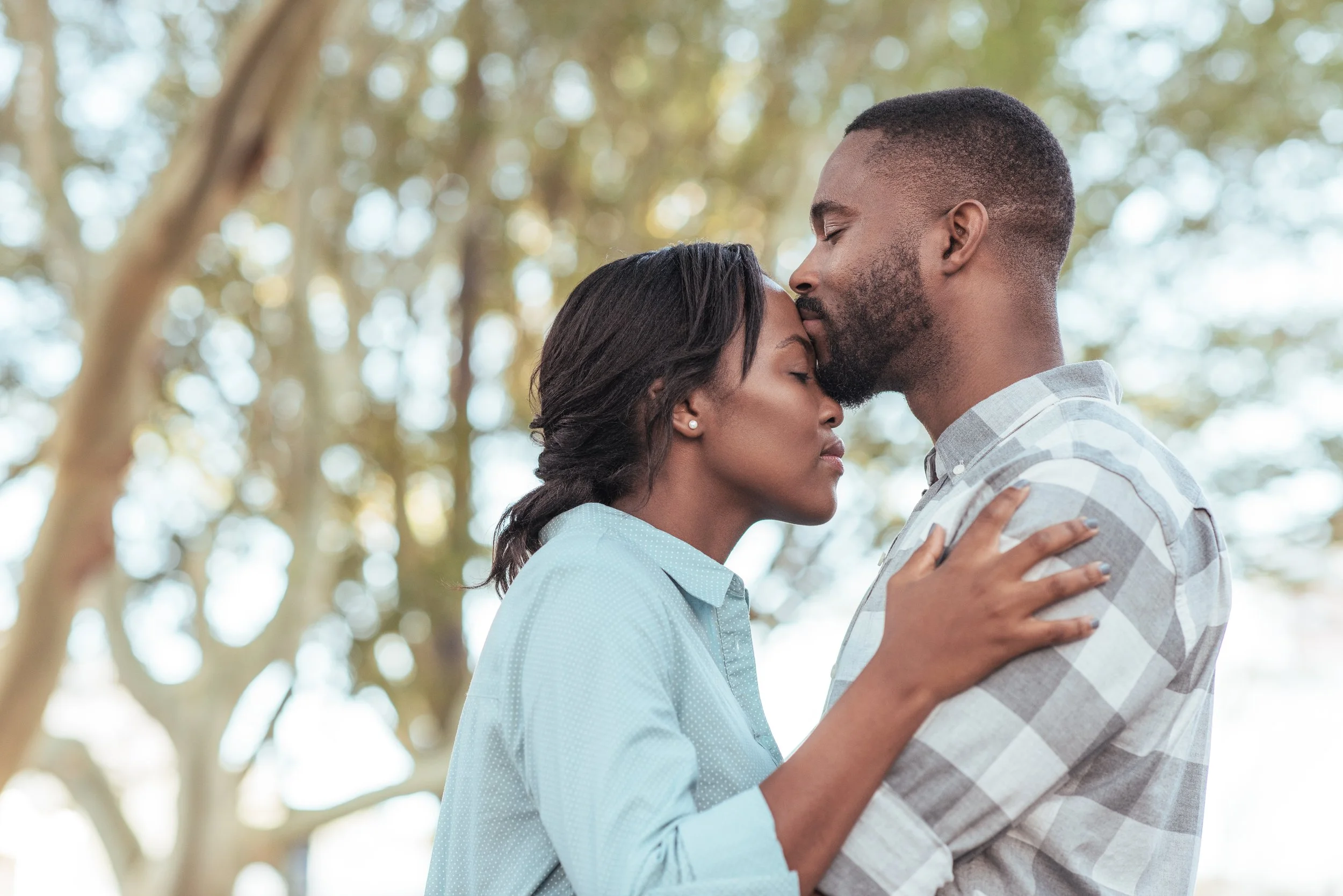 Couple embracing outdoors with trees in the background, eyes closed, and peaceful expressions.