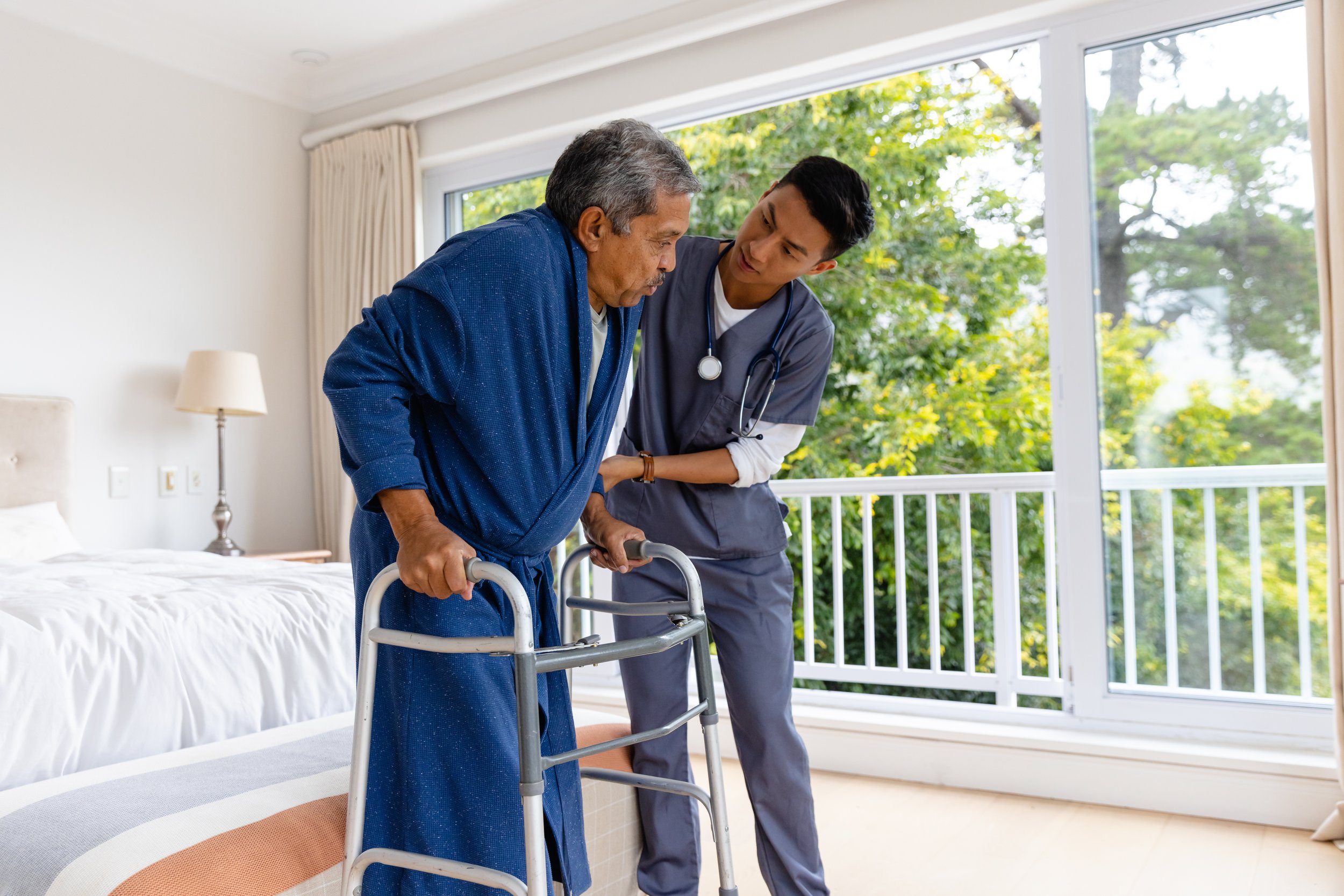 A healthcare professional assisting an elderly man using a walker in a bright room with large windows and greenery outside.