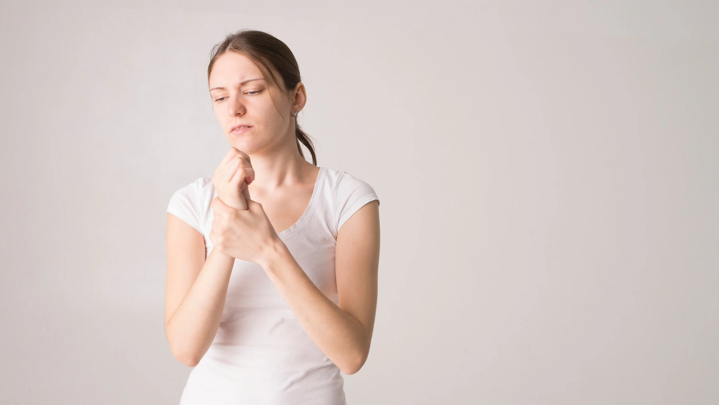 A young woman with brown hair in a ponytail, wearing a white t-shirt, looking concerned or thoughtful, holding her chin with her hand, standing against a plain light gray background.