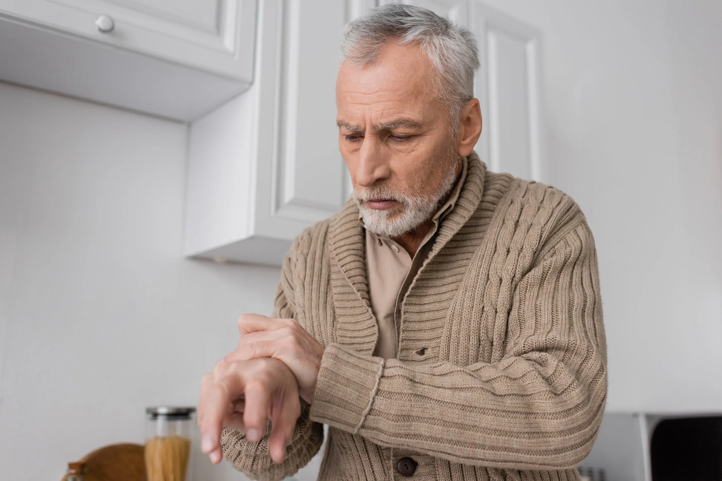 An elderly man with gray hair and a beard, wearing a beige cardigan sweater, looking at his wristwatch in a kitchen.