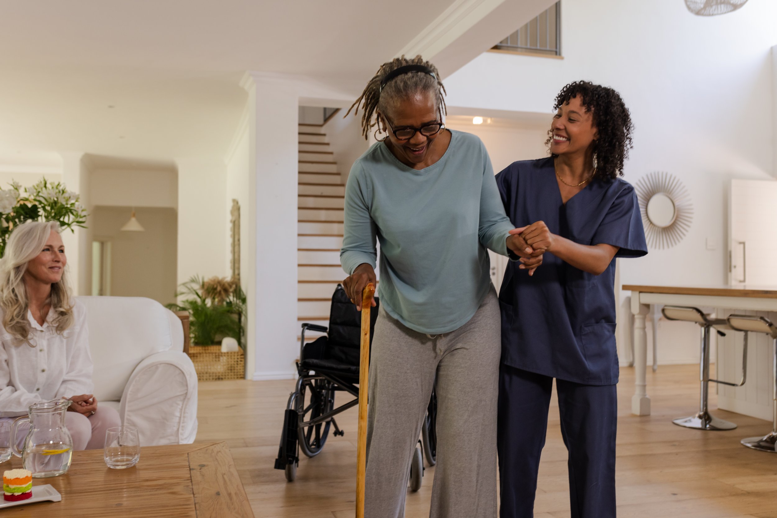 An elderly woman with a cane smiling as she is supported and comforted by a caregiver in a navy uniform in a bright, modern living room, with another woman sitting on a couch in the background.
