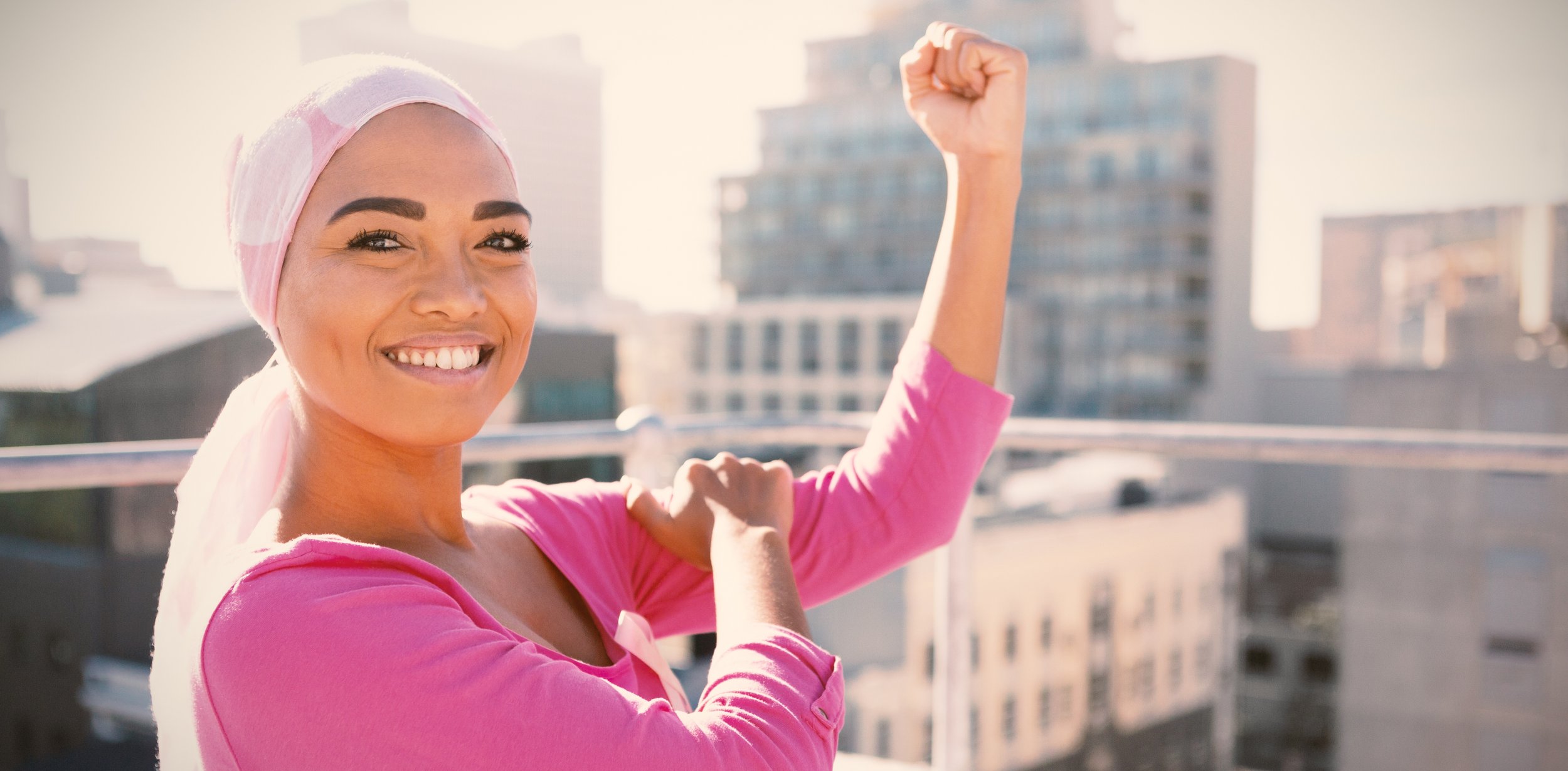 Happy woman wearing pink long sleeve shirt and a pink headscarf, flexing her arm on a rooftop with city buildings in the background.