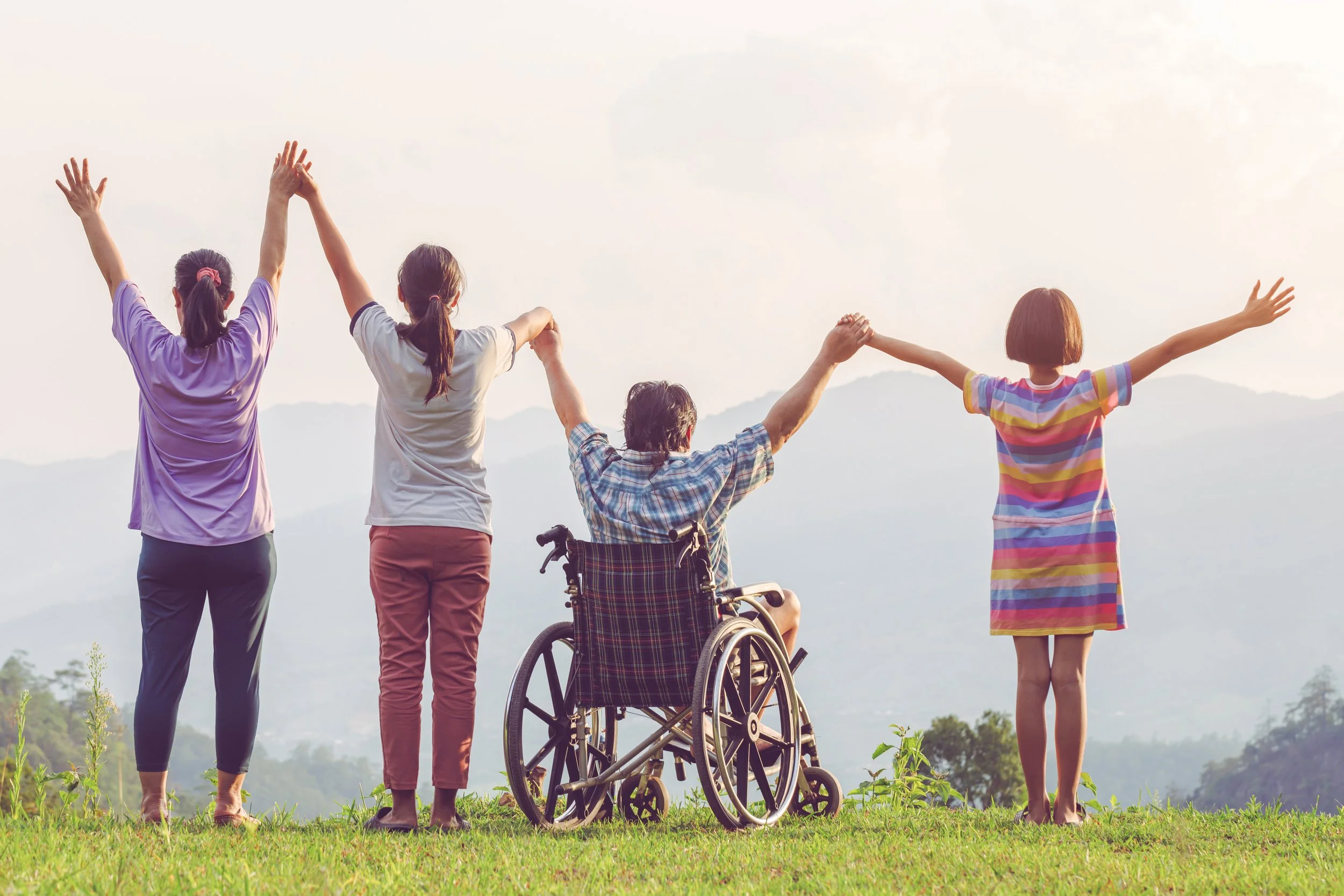 Four women holding hands and raising their arms, standing on grass with mountains in the background, one in a wheelchair, enjoying a scenic outdoor view.