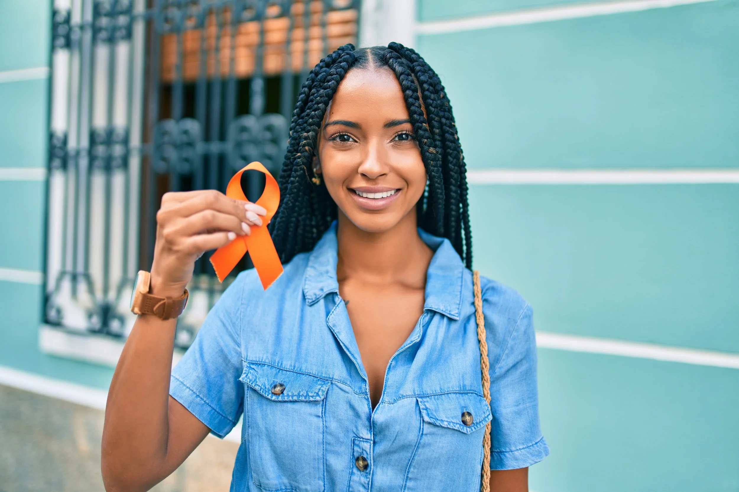 A woman with braided hair smiling and holding an orange awareness ribbon in front of a teal-colored wall.