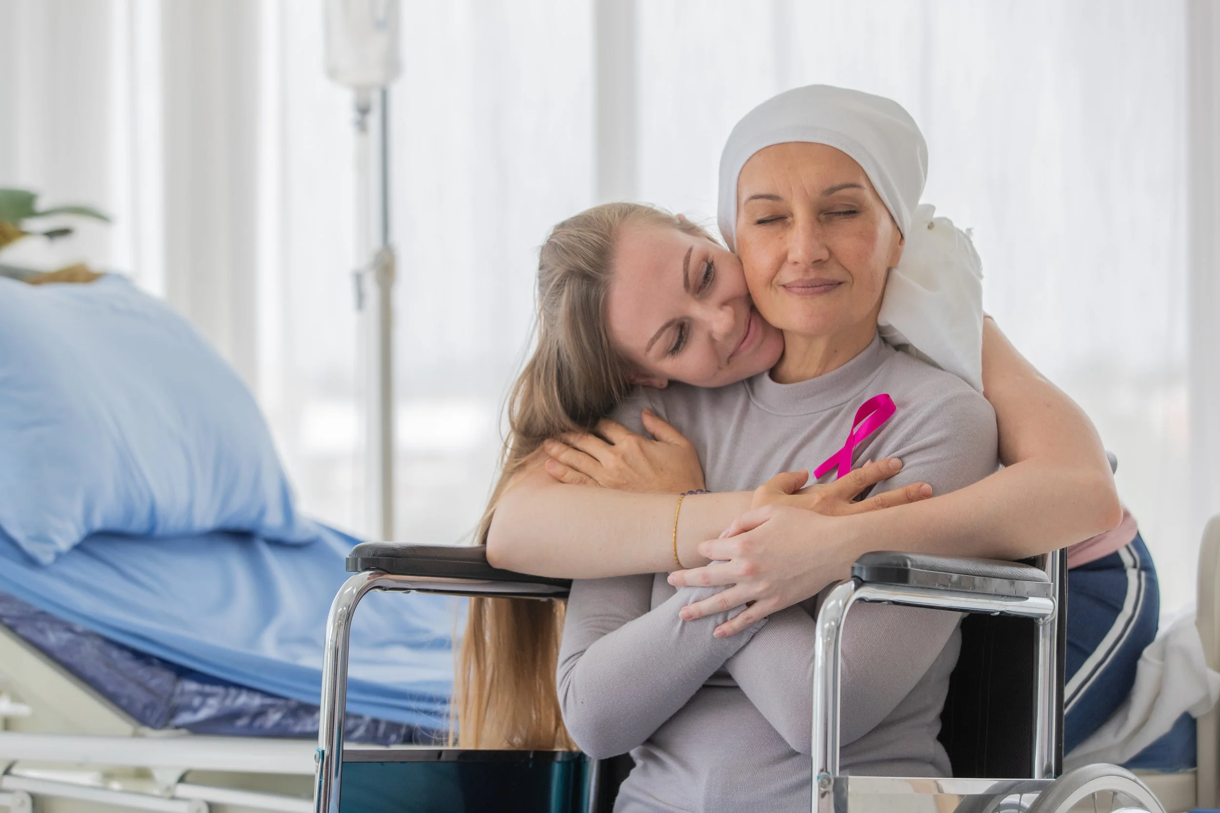 A woman in a wheelchair hugging a woman with a headscarf, both smiling and embracing in a hospital room.